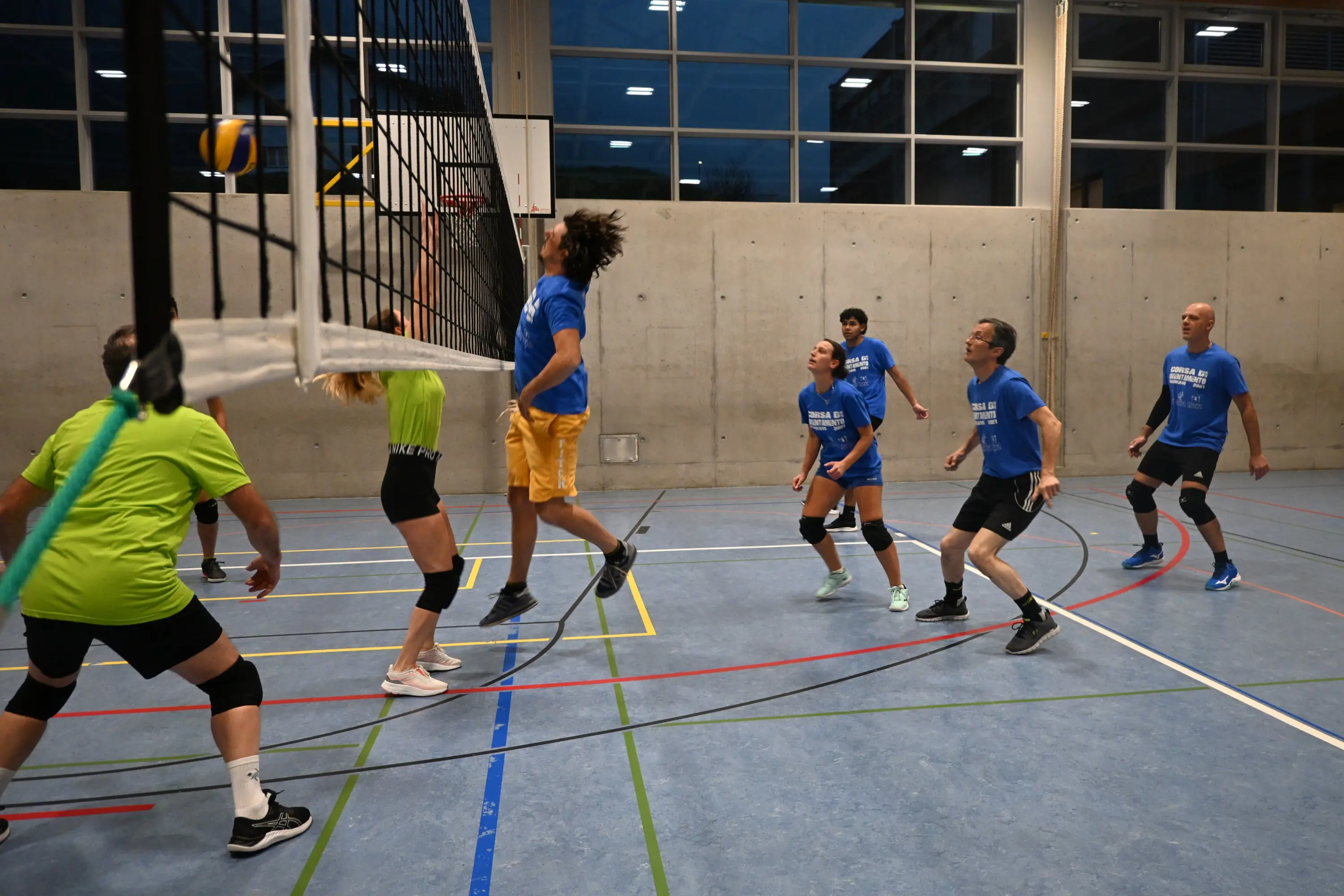 Una squadra di pallavolo in un campo al chiuso. Giocatori in magliette blu e verdi stanno per colpire una palla sopra la rete. Alcuni atleti sono in movimento, mentre altri osservano attentamente. L'illuminazione è artificiale e il