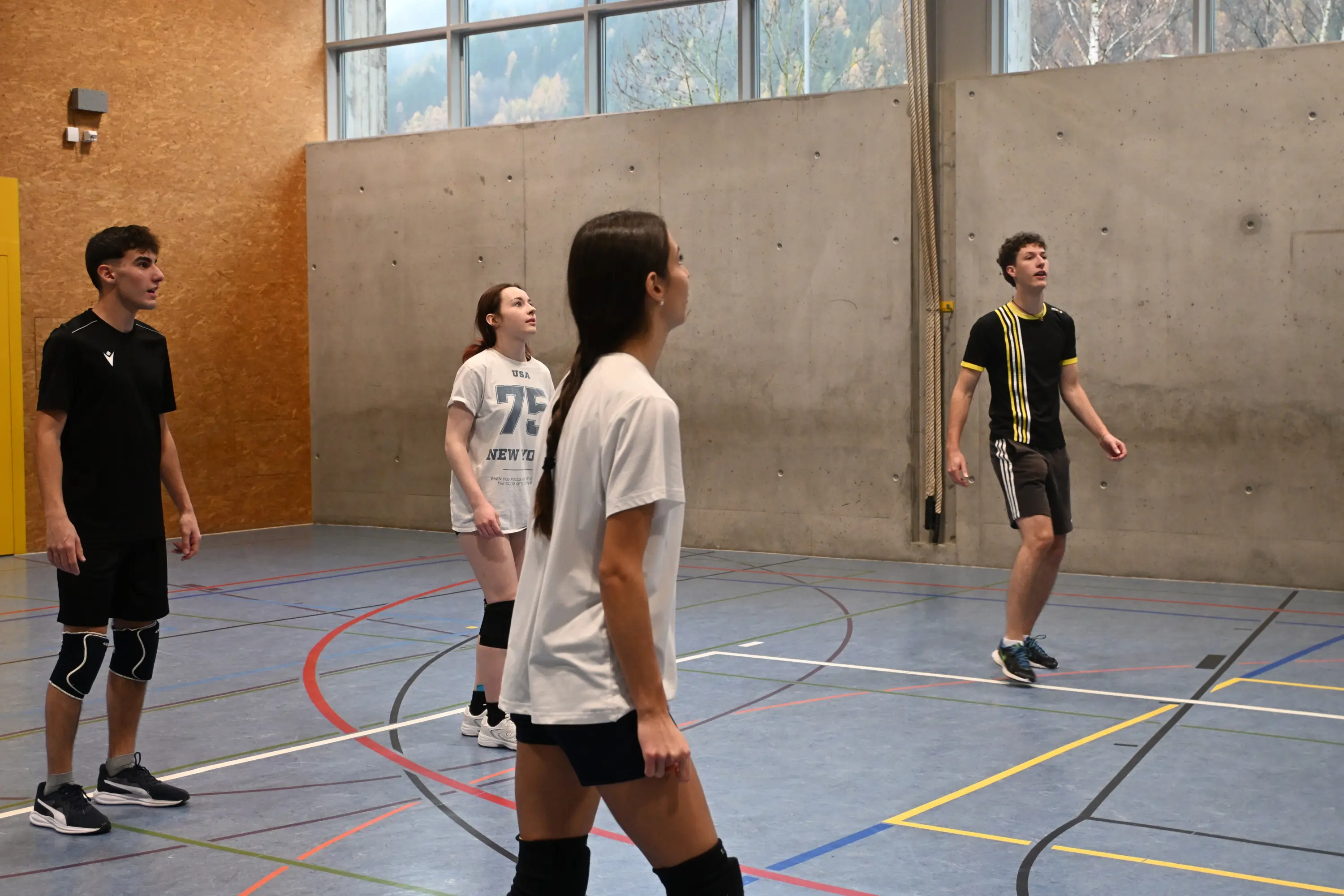 Quattro giovani atleti in una palestra, pronti a giocare a pallavolo. Due ragazzi e due ragazze sono in posizione di attesa, osservando la palla. Sullo sfondo si vede una parete di cemento e finestre che lasciano filtrare la luce