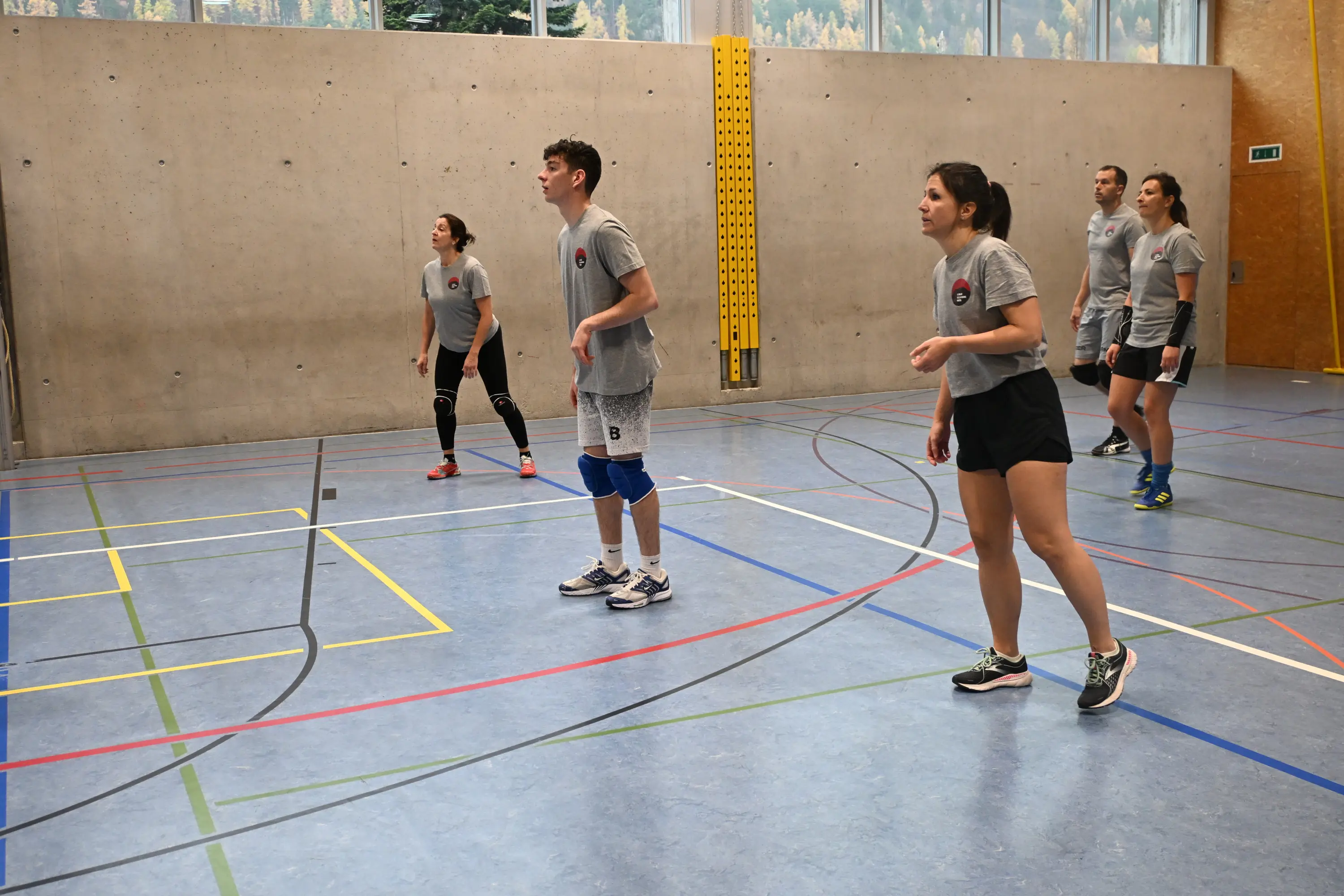 Un gruppo di giovani atleti si prepara a ricevere una palla in un campo da pallavolo. Indossano magliette grigie e pantaloni sportivi, con espressioni concentrate. Sullo sfondo, si intravedono le pareti di una palestra.