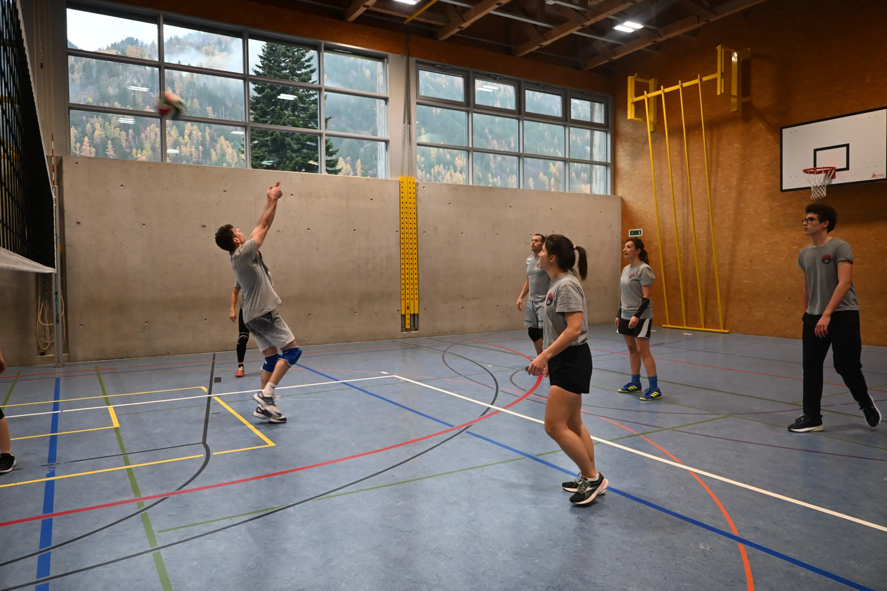 Giovani atleti si allenano in una palestra, con un giocatore che esegue un servizio di pallavolo mentre gli altri osservano. Sfondo di finestre con vista su alberi e montagne.