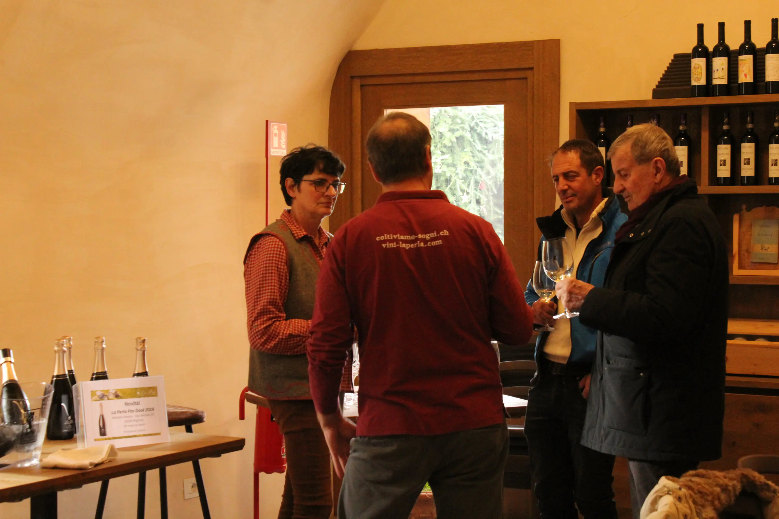 Quattro persone in un ambiente rustico, mentre degustano vini. Uno dei partecipanti indossa una maglietta rossa con il logo di un'azienda vinicola. Sullo sf