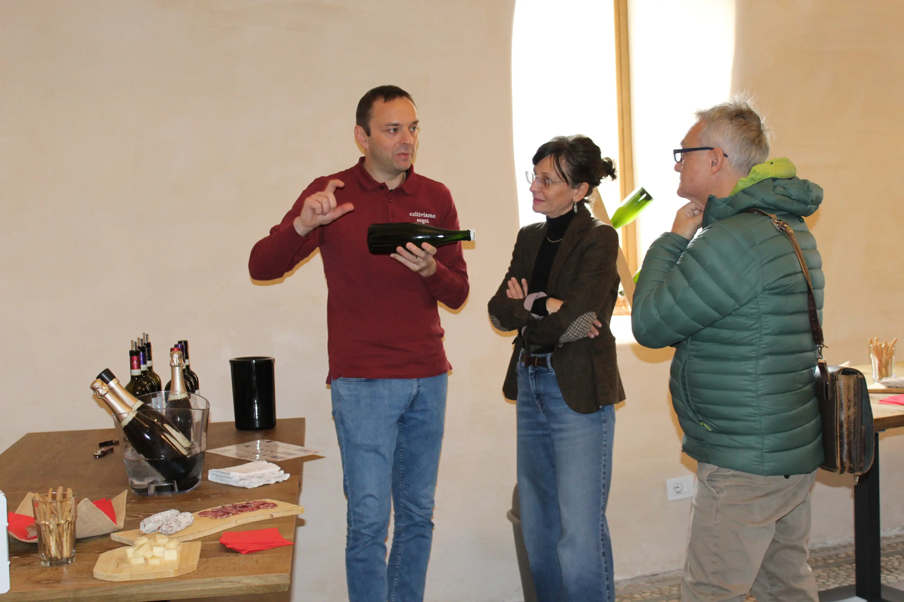 Un uomo spiega qualcosa mentre tiene una bottiglia, accompagnato da due persone che ascoltano attentamente. Su un tavolo ci sono bottiglie di vino e alcuni formaggi. La scena