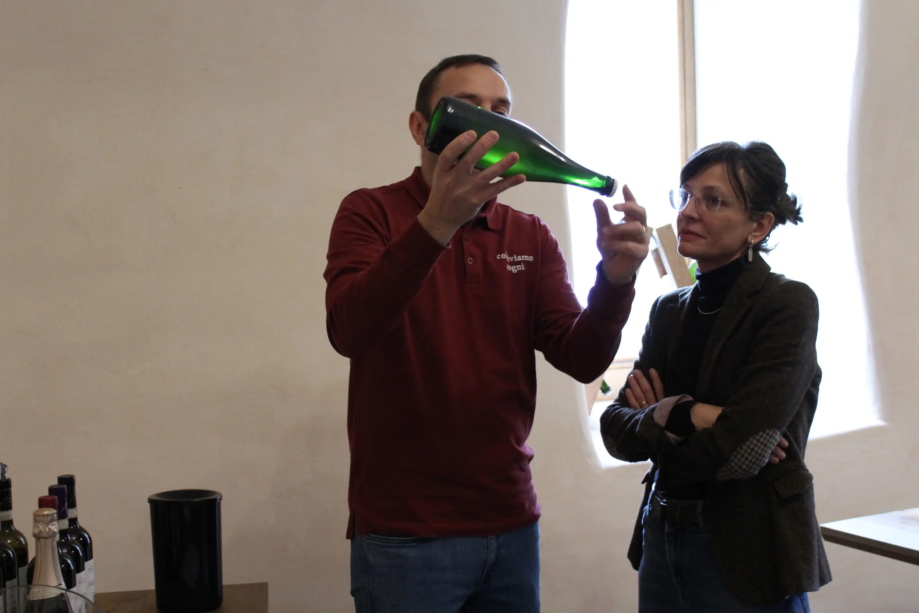 Un uomo tiene una bottiglia di vino verde, spiegando qualcosa a una donna che lo ascolta attentamente, con le braccia incrociate. Sullo sfondo si vedono alcune