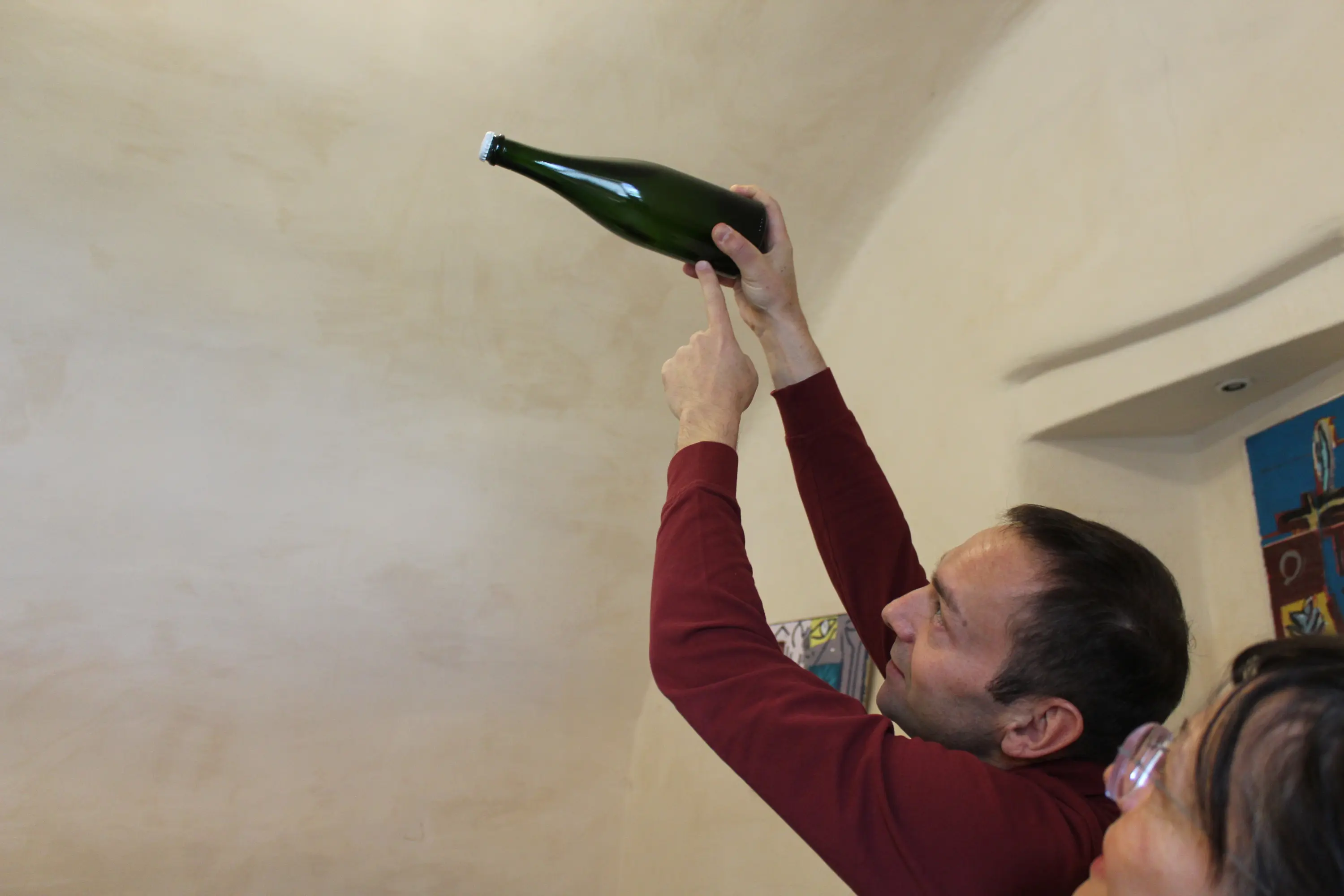 Una persona alza una bottiglia verde contro un soffitto, mentre un'altra persona osserva con interesse. La parete è di un colore neutro.