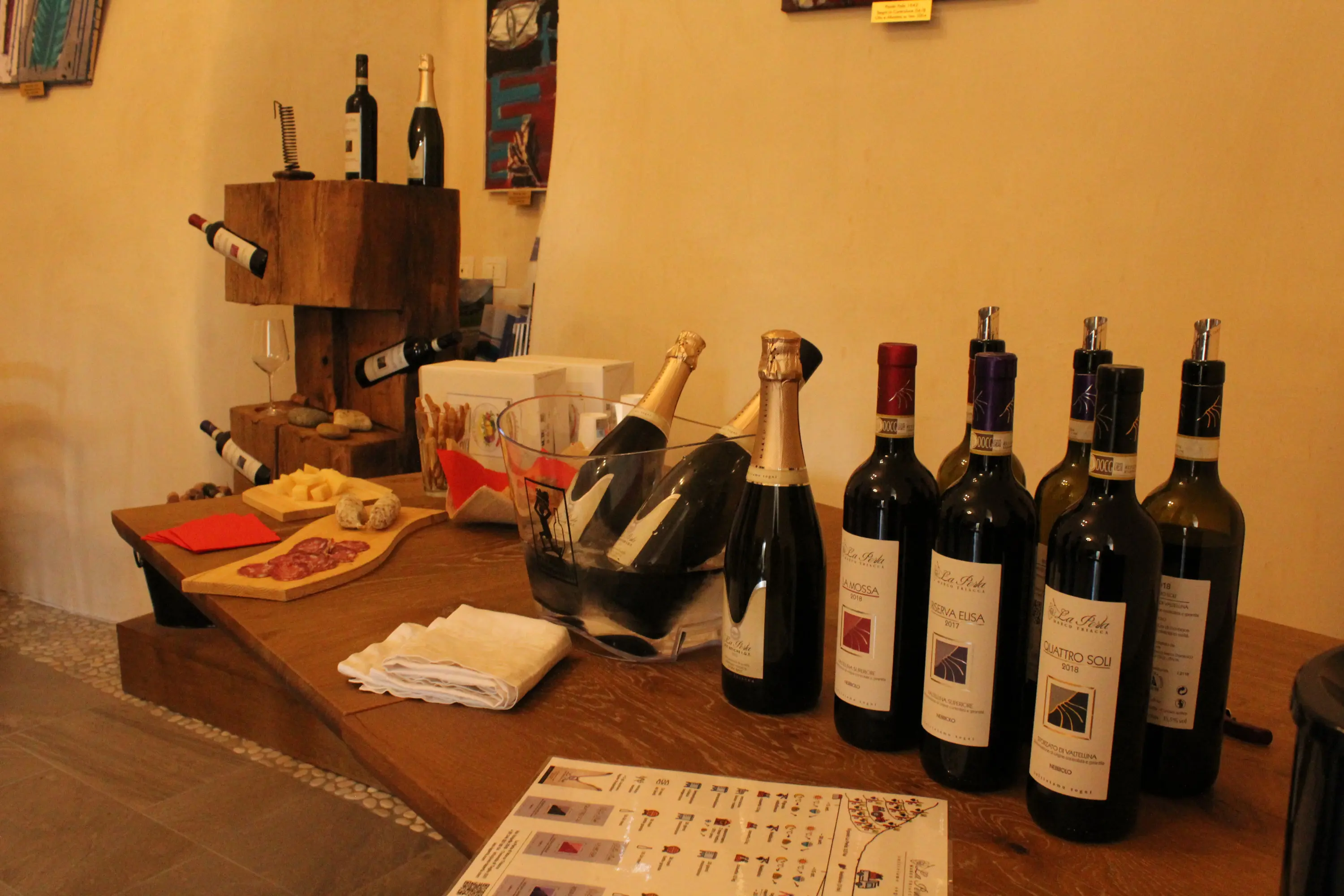 Una tavola in legno con diverse bottiglie di vino, alcune con etichette colorate, un secchiello con spumante e taglieri di salumi. Sullo sf