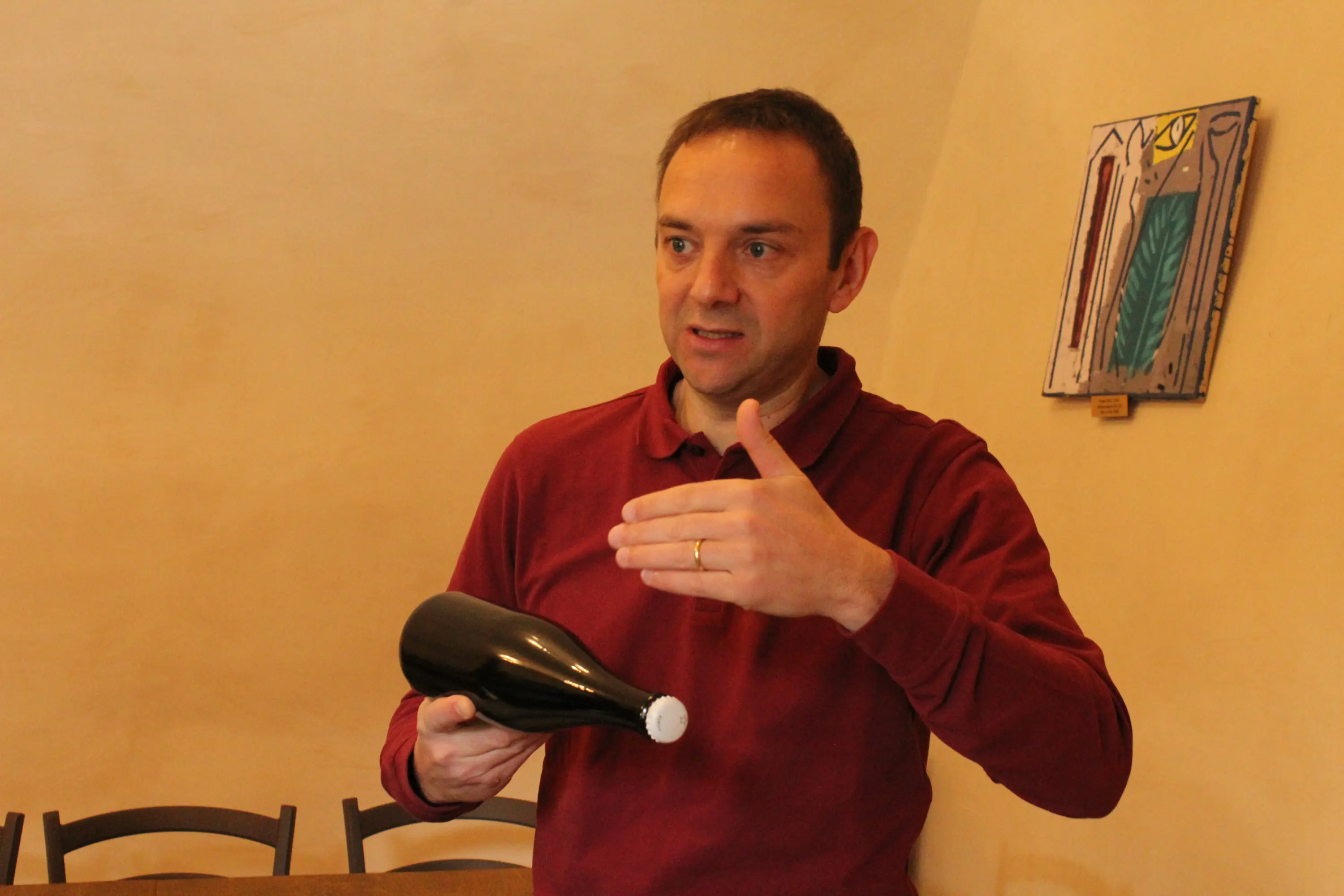 Un uomo in maglione rosso tiene una bottiglia scura e spiega qualcosa, con un gesto della mano. Sullo sfondo si intravede un'opera d'arte app