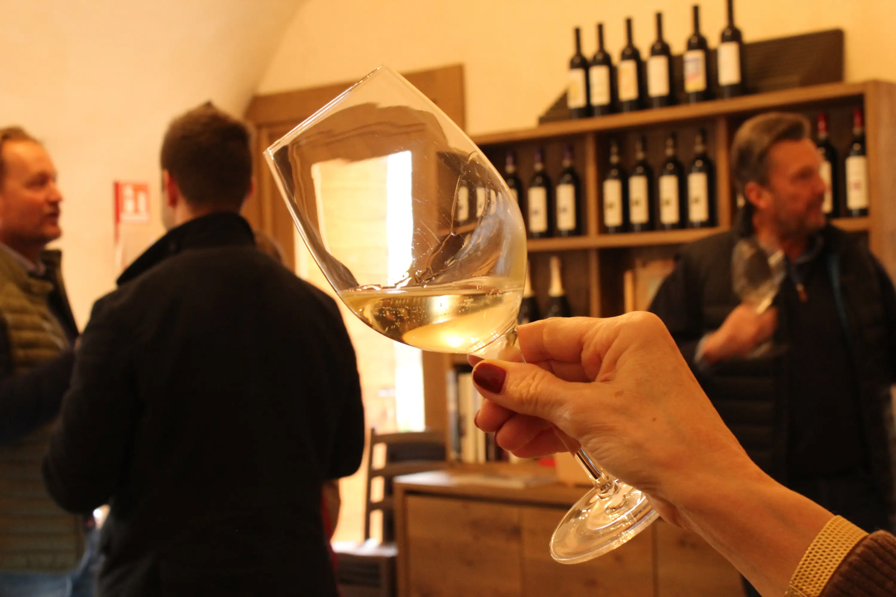 Un bicchiere di vino bianco viene sollevato verso la fotocamera con delle persone sullo sfondo, mentre si trovano in una cantina. Sullo sfondo ci sono