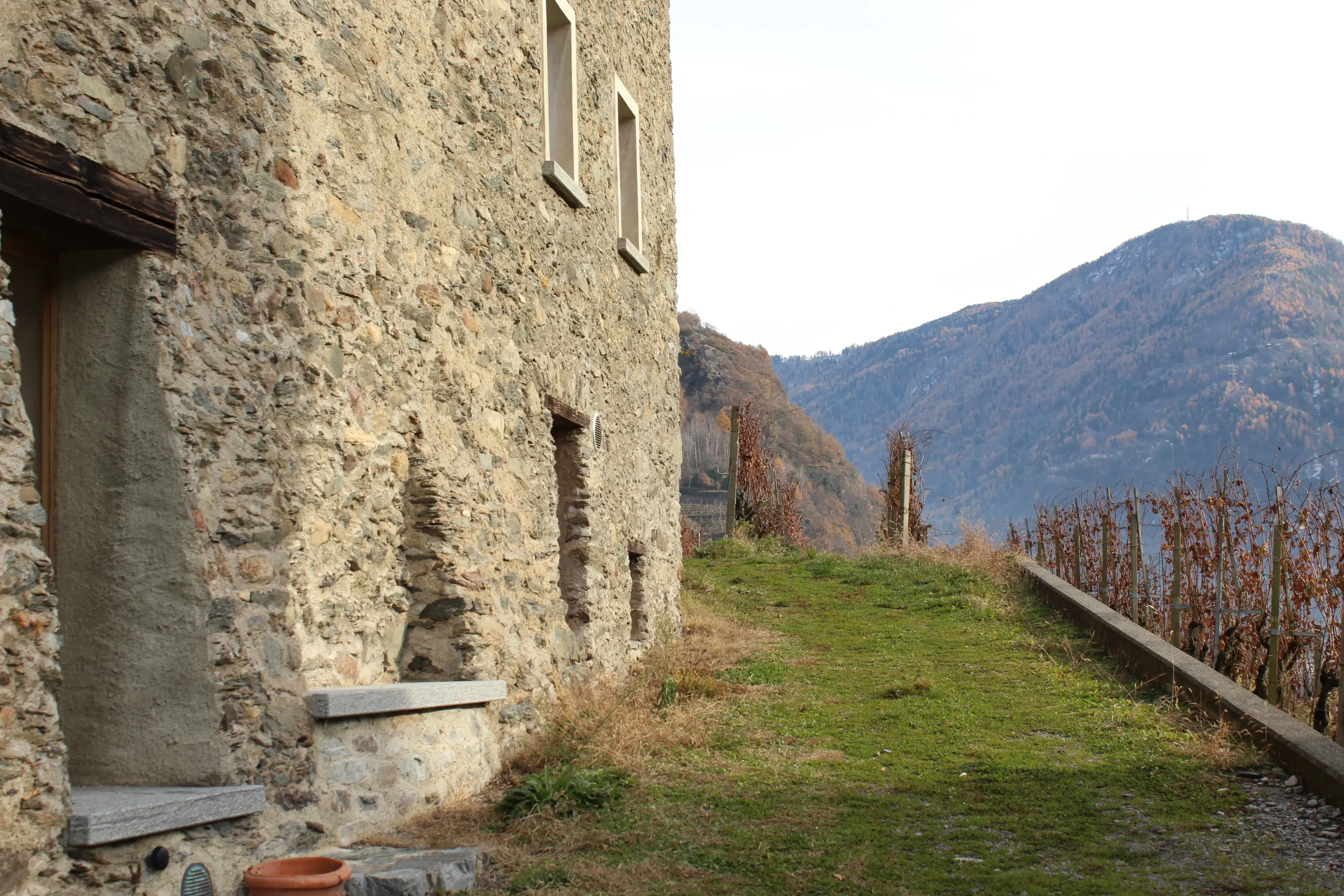 Un vecchio edificio di pietra con mura rustiche, situato lungo un sentiero che si snoda attraverso un paesaggio montano. Sullo sfondo si vedono colline verdi