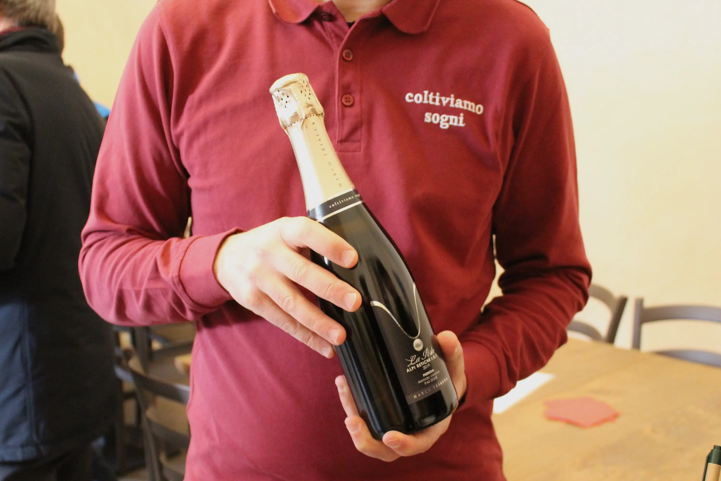 Un uomo tiene in mano una bottiglia di spumante nera con etichetta elegante, indossando una maglietta rossa con la scritta "coltiviamo sogni