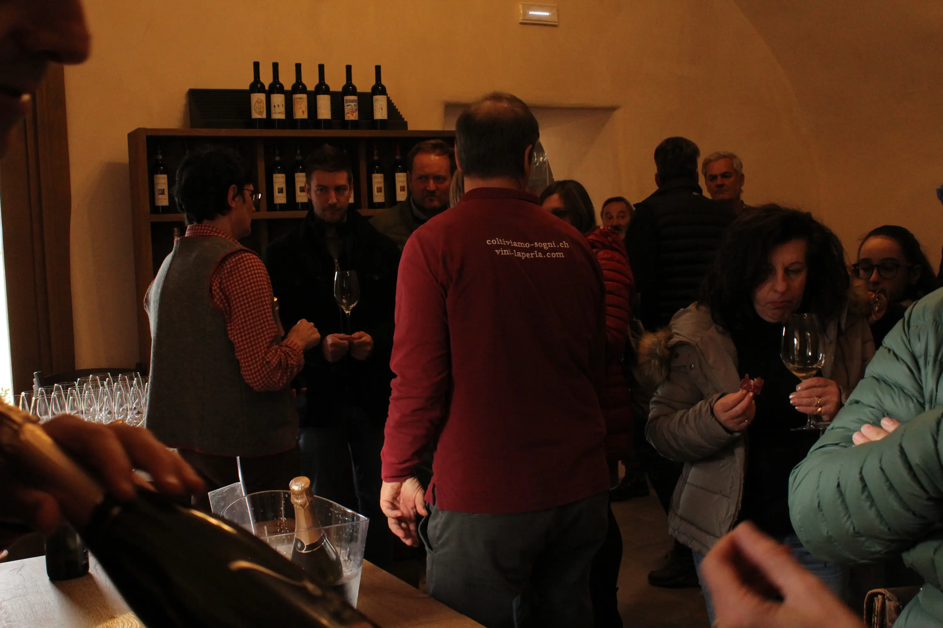 Una cantina affollata, con persone che degustano vino e chiacchierano. Bottiglie di vino sono visibili sugli scaffali. Alcuni ospiti tengono bicch