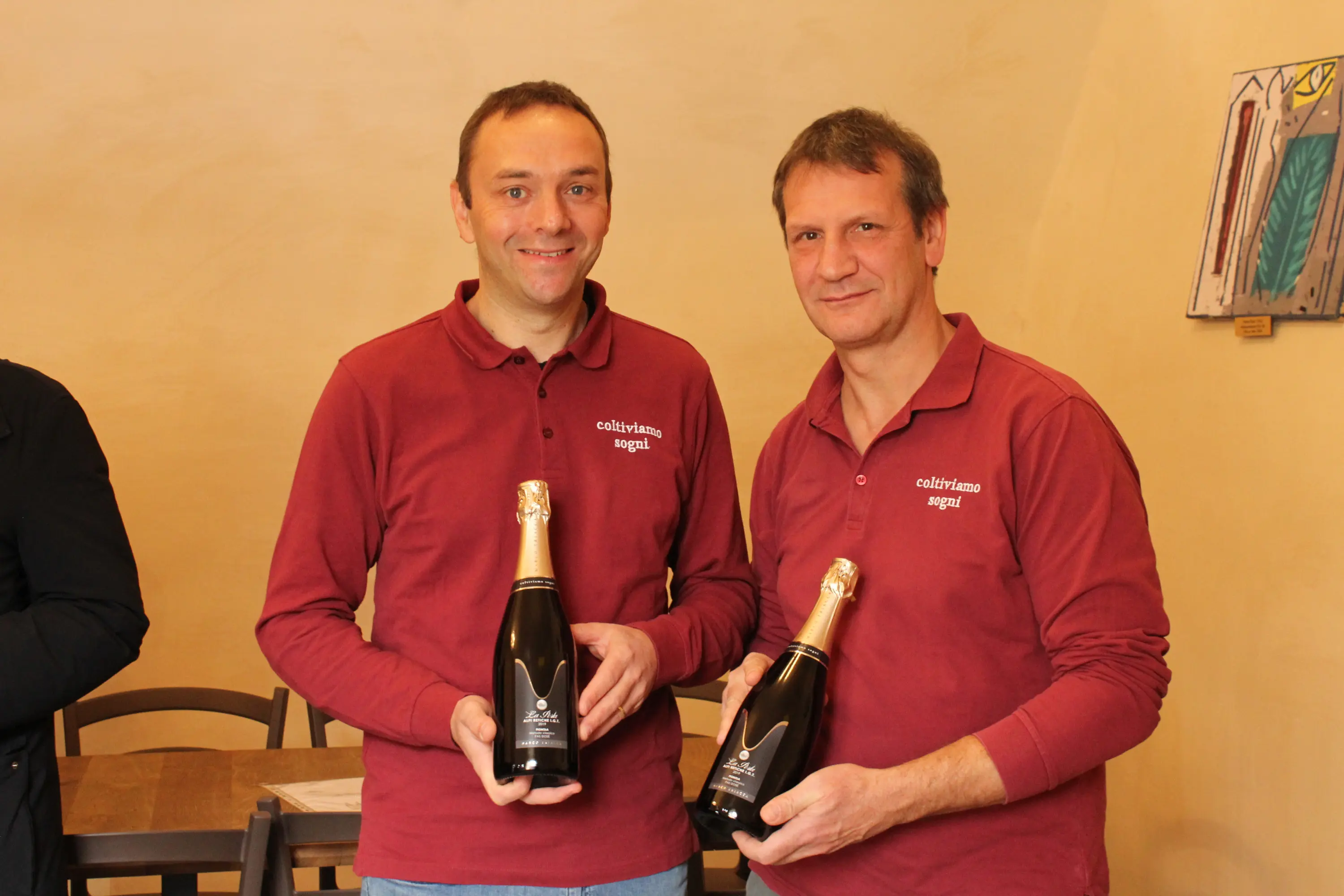 Due uomini sorridenti indossano magliette rosse con il logo "caliciamo segui" e mostrano bottiglie di champagne. Sullo sfondo, un ambiente semplice e acc