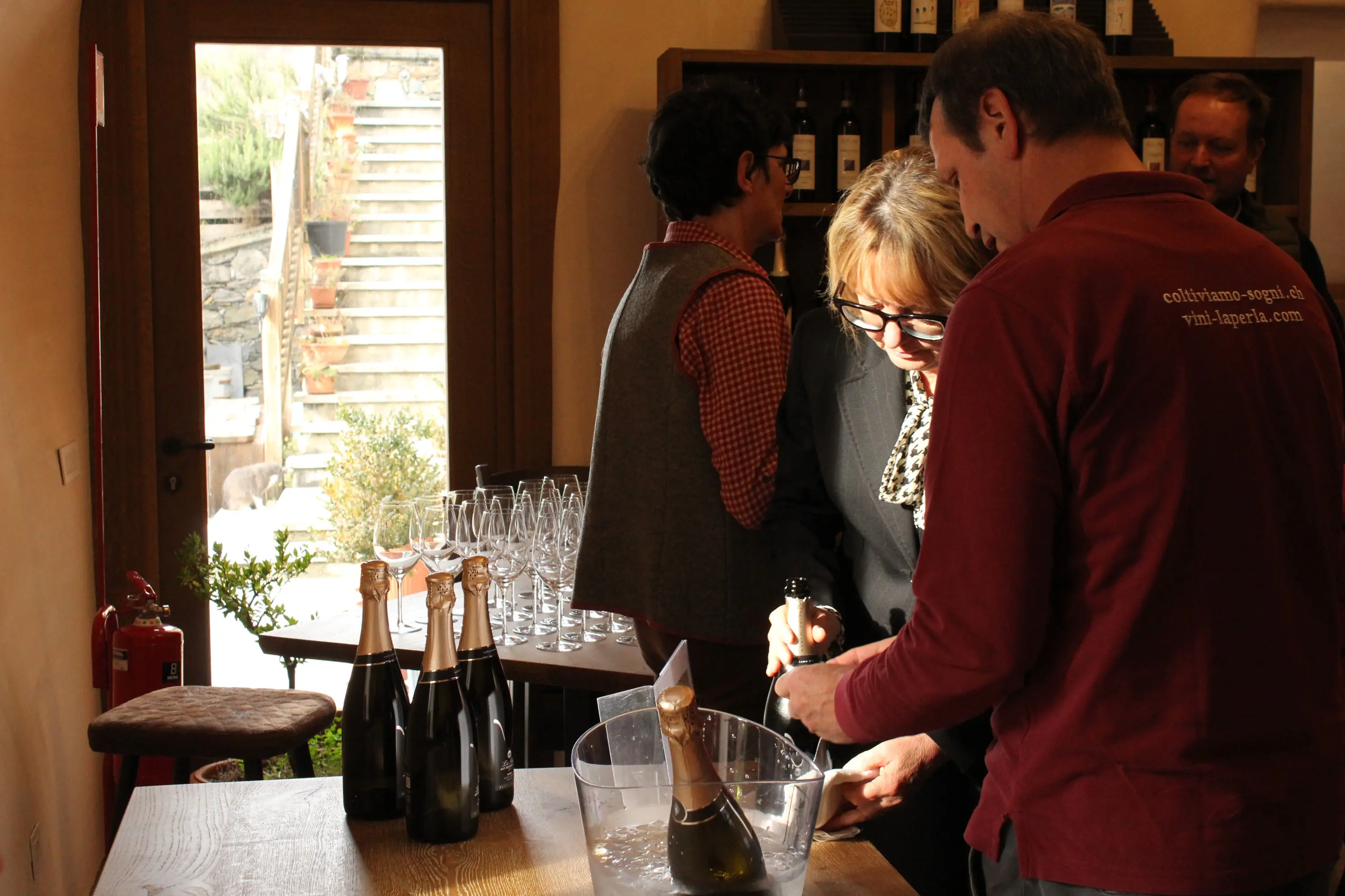 Una degustazione di vini in una cantina, con un uomo che stappa una bottiglia di spumante mentre una donna lo osserva. Sullo sfondo, si vedono altre persone