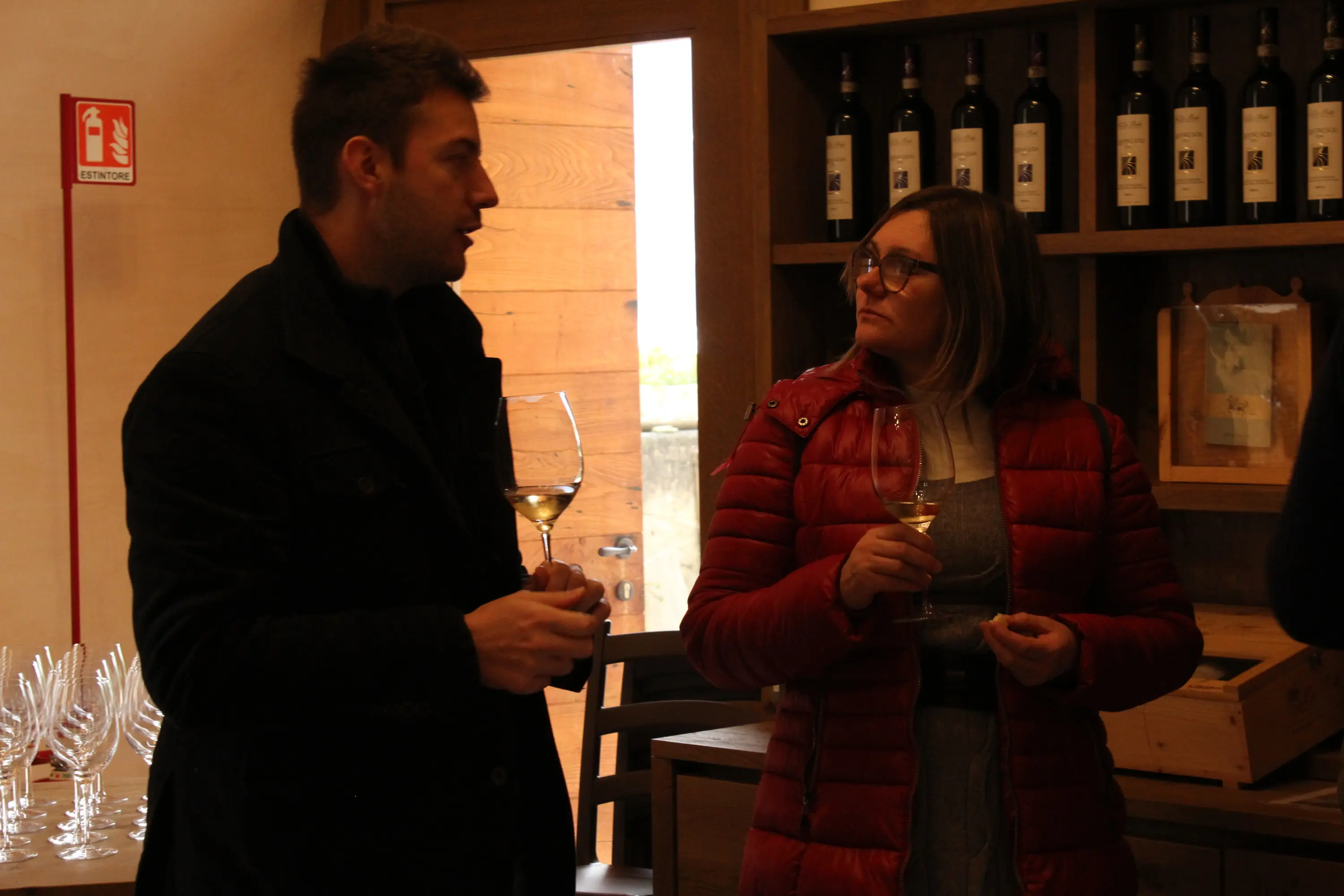 Due persone conversano all'interno di una cantina, ognuna con un bicchiere di vino. Una donna indossa una giacca rossa e guarda l'uomo, che ind