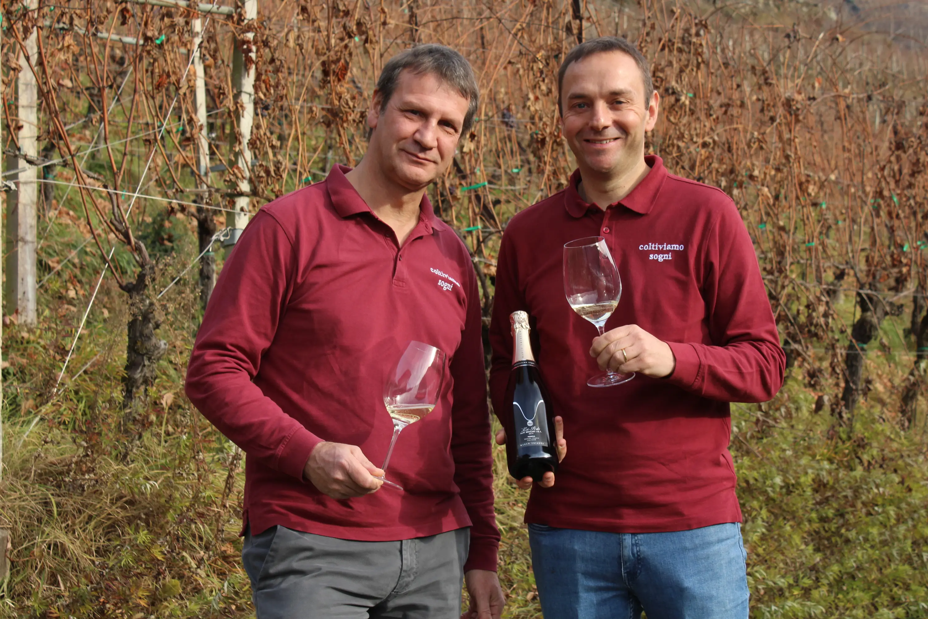 Due uomini sorridenti in un vigneto, ciascuno tiene un bicchiere di vino e una bottiglia. Indossano magliette rosse con il logo di un'azienda vin