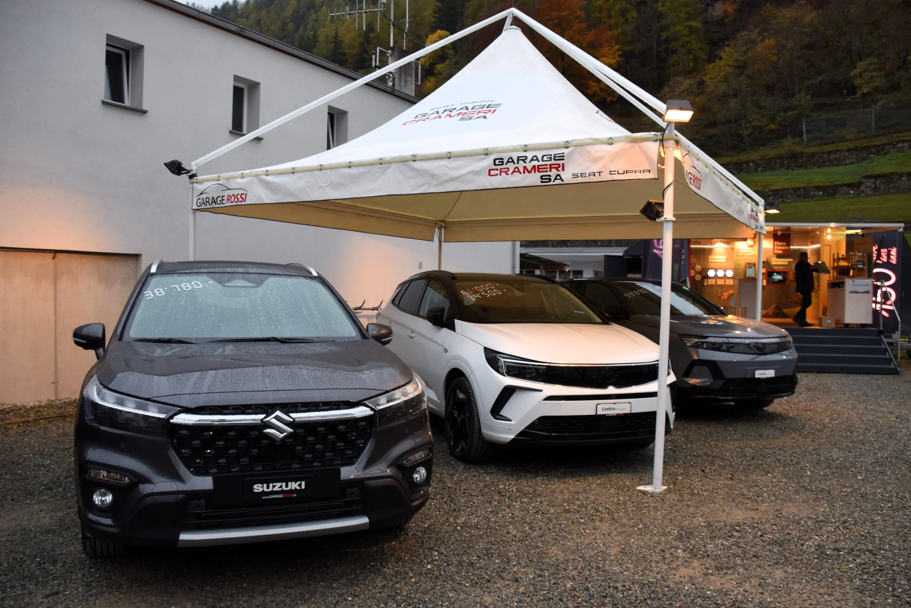 Tre auto esposte sotto un tendone pubblicitario. Due SUV, uno nero e uno bianco, affiancati a un SUV grigio scuro, con un'insegna di un concessionario di auto visibile in background.