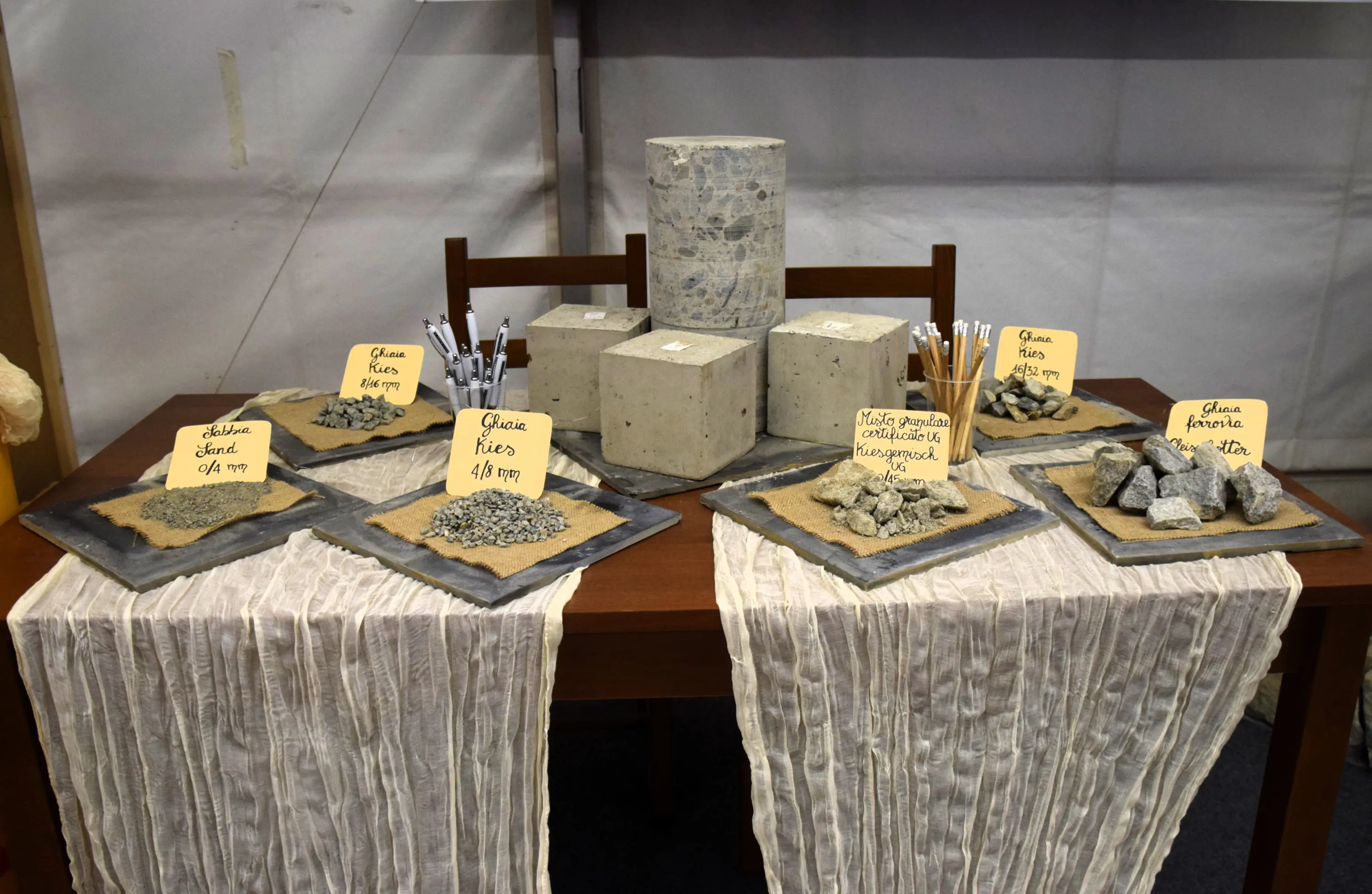 Un tavolo espositivo con diverse tipologie di materiali, tra cui campioni di cemento e pietre, disposti su piatti decorativi. Cartelli informativi descrivono ciascun campione. Un cilindro di cemento si trova al centro della disposizione.