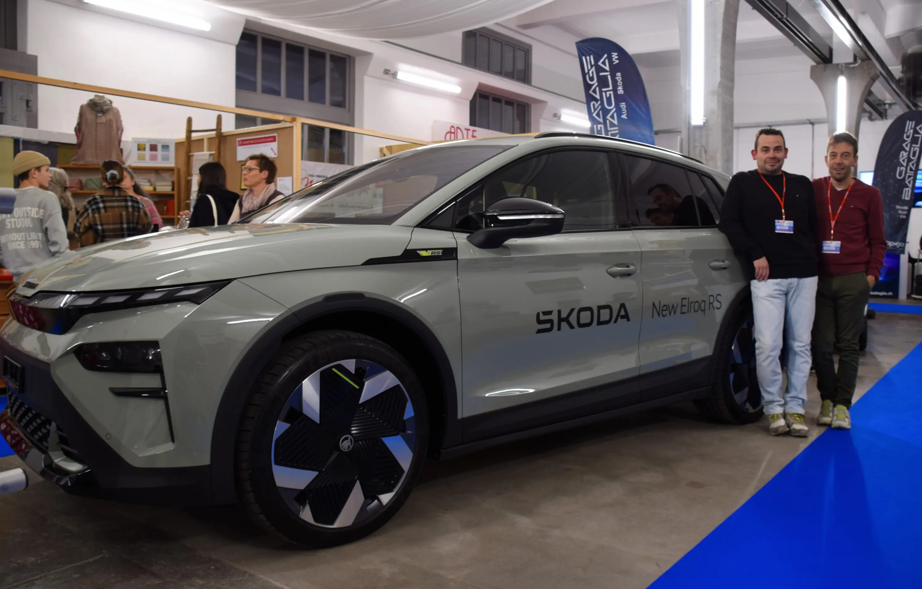 Due uomini sorridono accanto a un'auto elettrica Skoda New Etrusco, con un design moderno e dettagli distintivi, esposta in un ambiente fieristico.