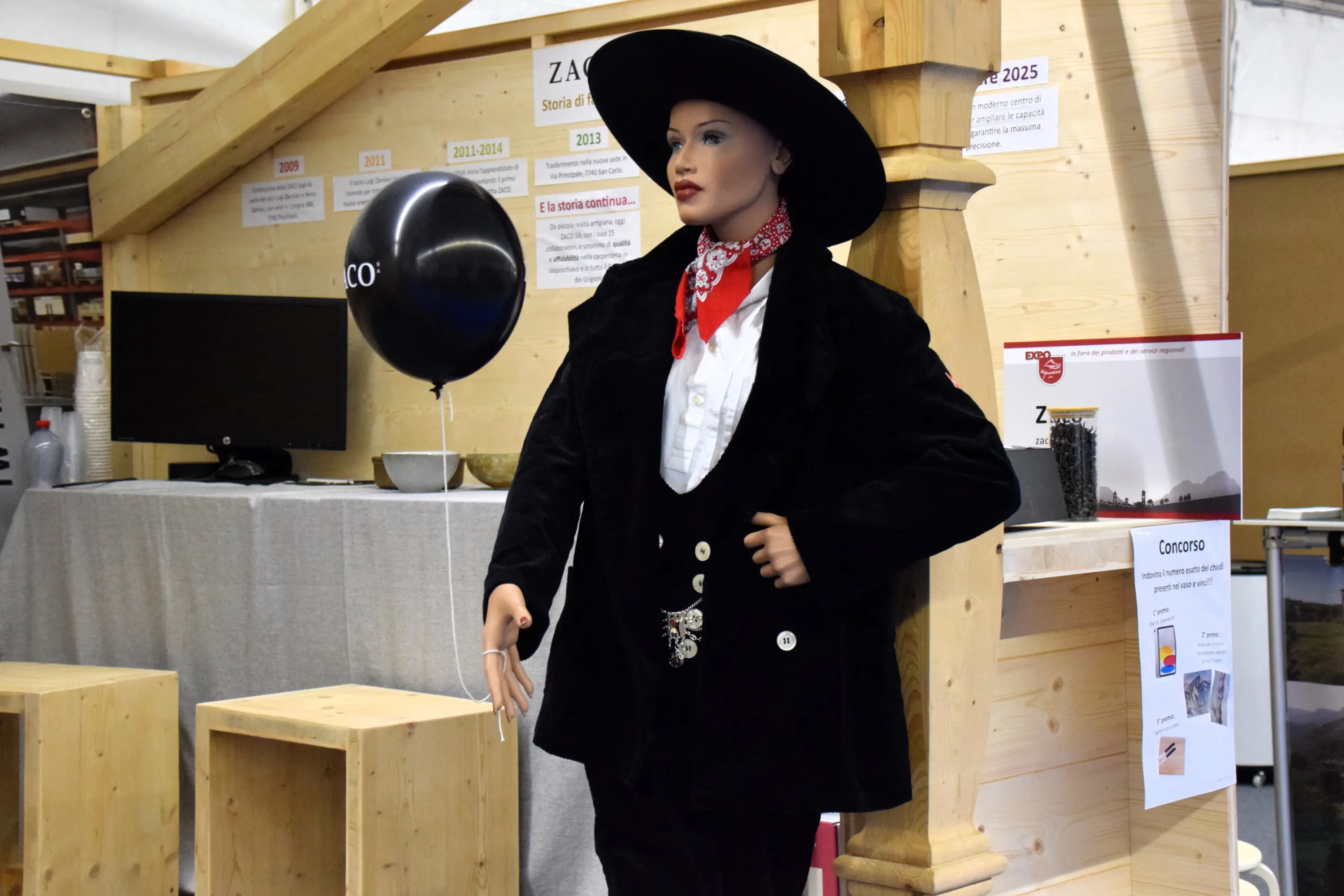 Un manichino vestito con un abito in stile cowboy, completo di cappello e bandana rossa, si trova all'interno di uno spazio espositivo. Sullo sfondo ci sono tavoli di legno e un palloncino nero.