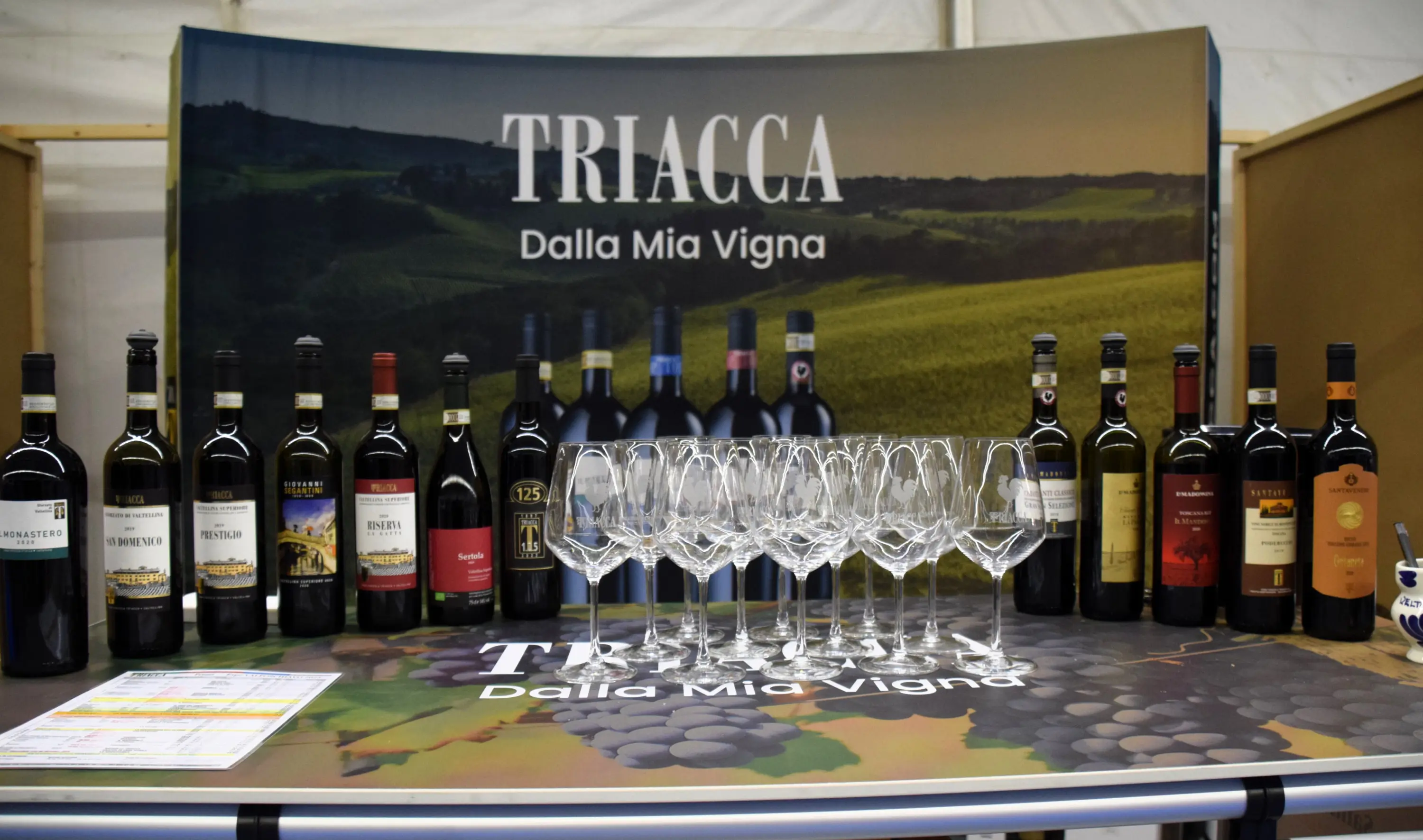 Una tavola con bottiglie di vino disposte in fila, accompagnate da bicchieri di vetro trasparenti. Sullo sfondo, un grande banner che mostra il nome "Triacca" e il paesaggio delle vigne.