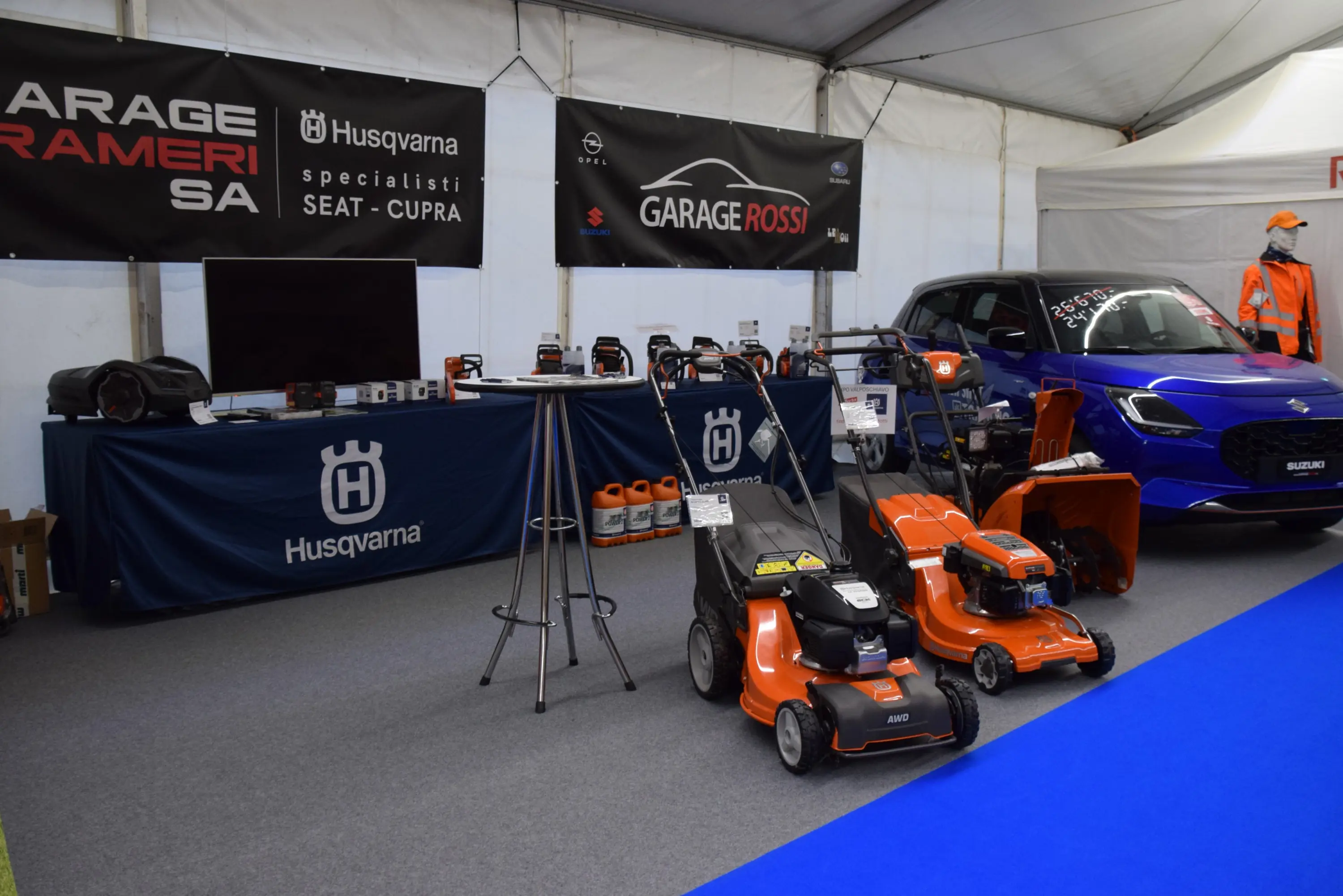 Spazio espositivo con attrezzature da giardino Husqvarna, tra cui tosaerba. Sullo sfondo, un'auto blu e insegne di concessionari di auto.