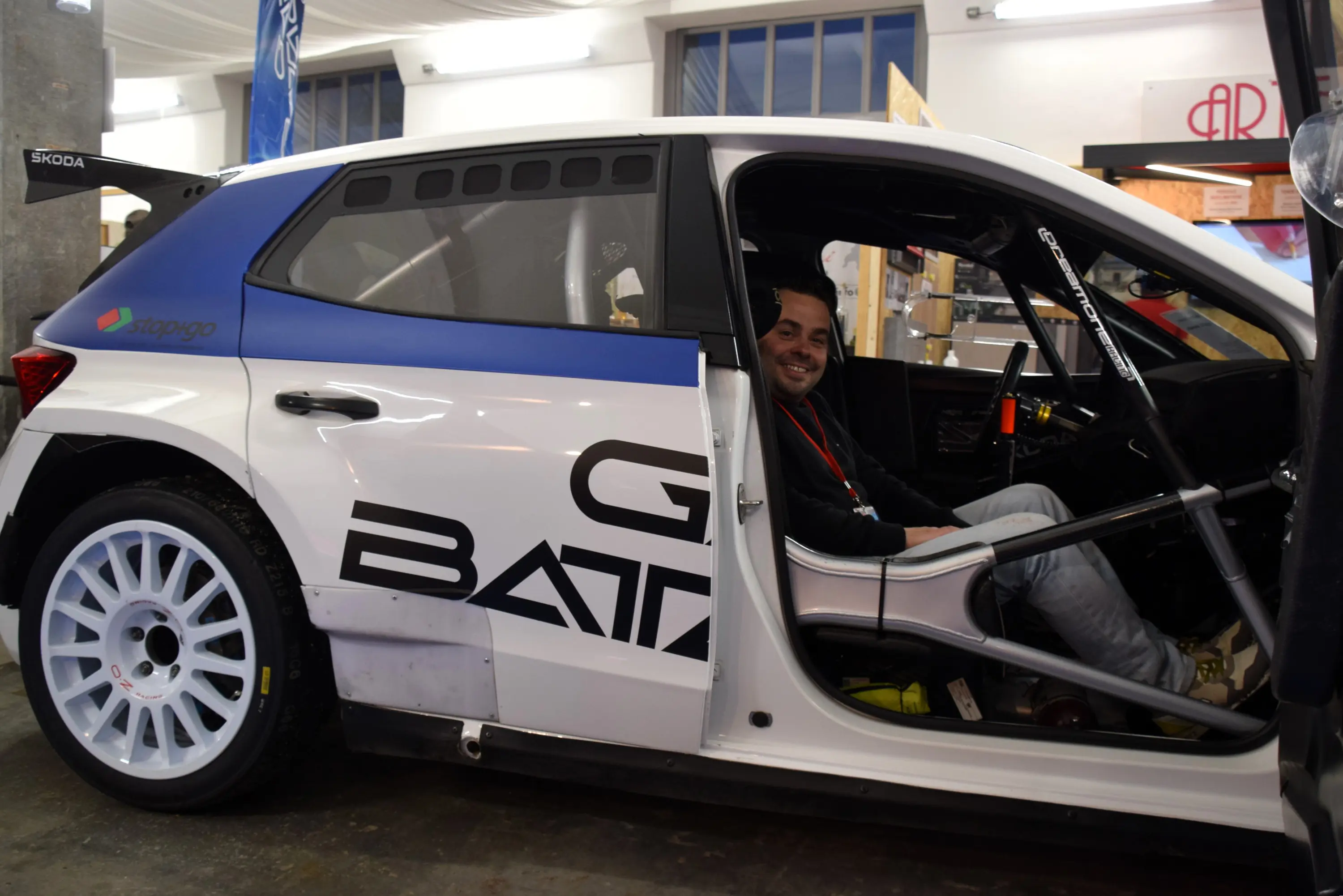 Un uomo sorridente seduto all'interno di una auto da rally con livrea bianca e blu. Il veicolo presenta sponsor e dettagli sportivi.