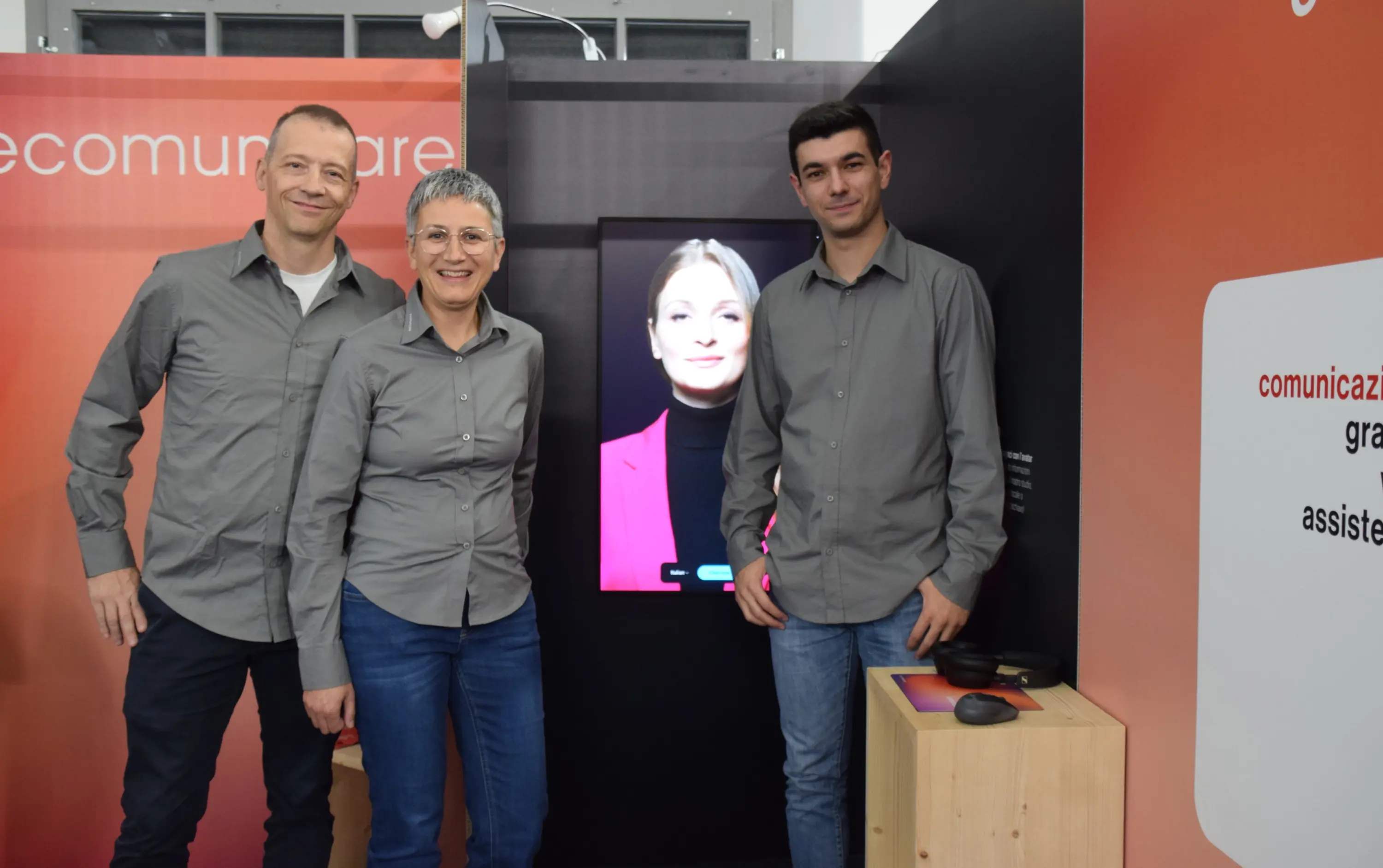 Tre persone sorridenti in abiti grigi posano davanti a uno schermo che mostra un'immagine di una donna con capelli lunghi e un blazer rosso. Sullo sfondo ci sono elementi minimalisti di design.