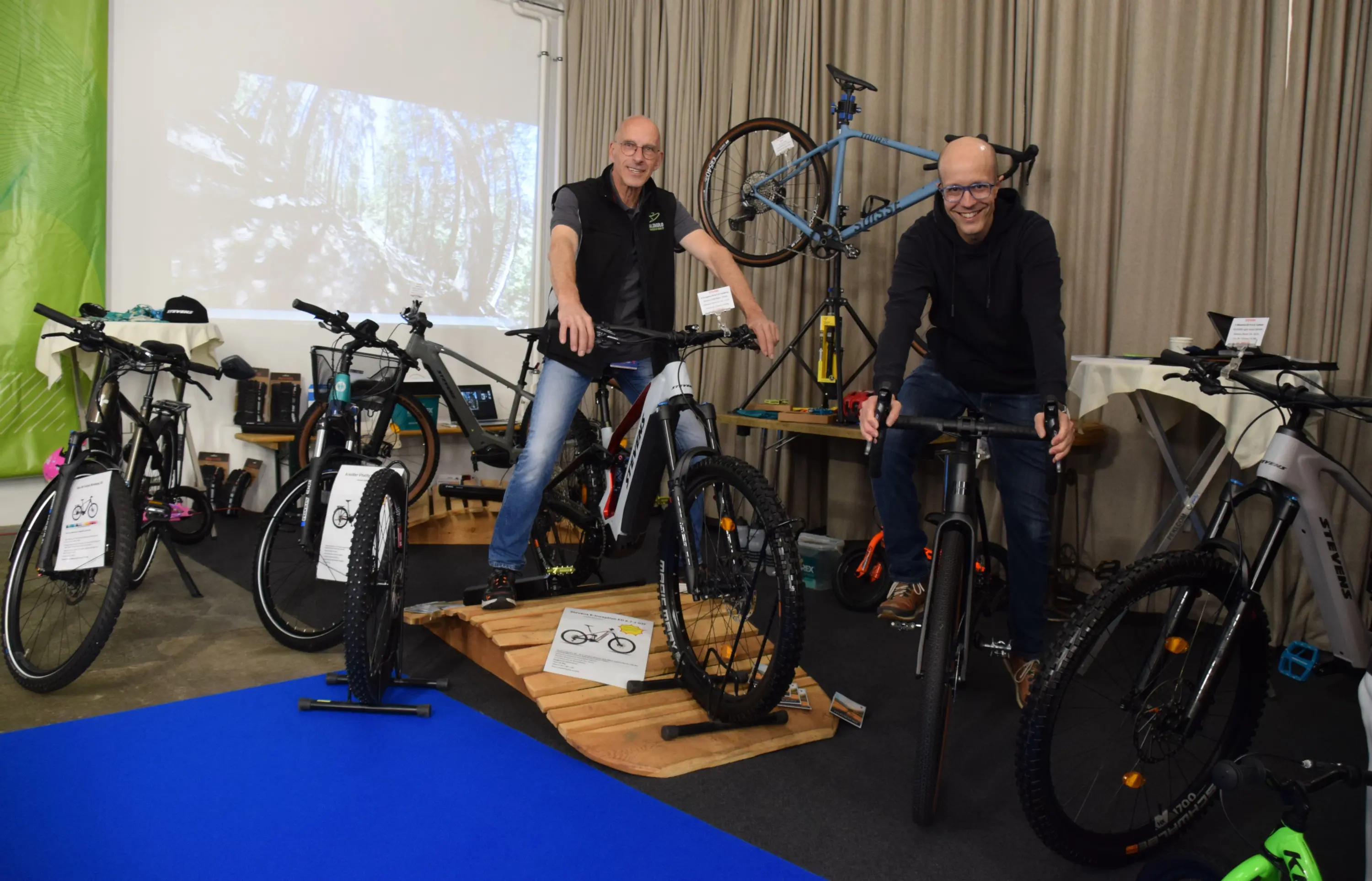 Due uomini sorridenti posano accanto a diverse biciclette, in un ambiente fieristico con un pavimento blu e un fondo di tende grigie.