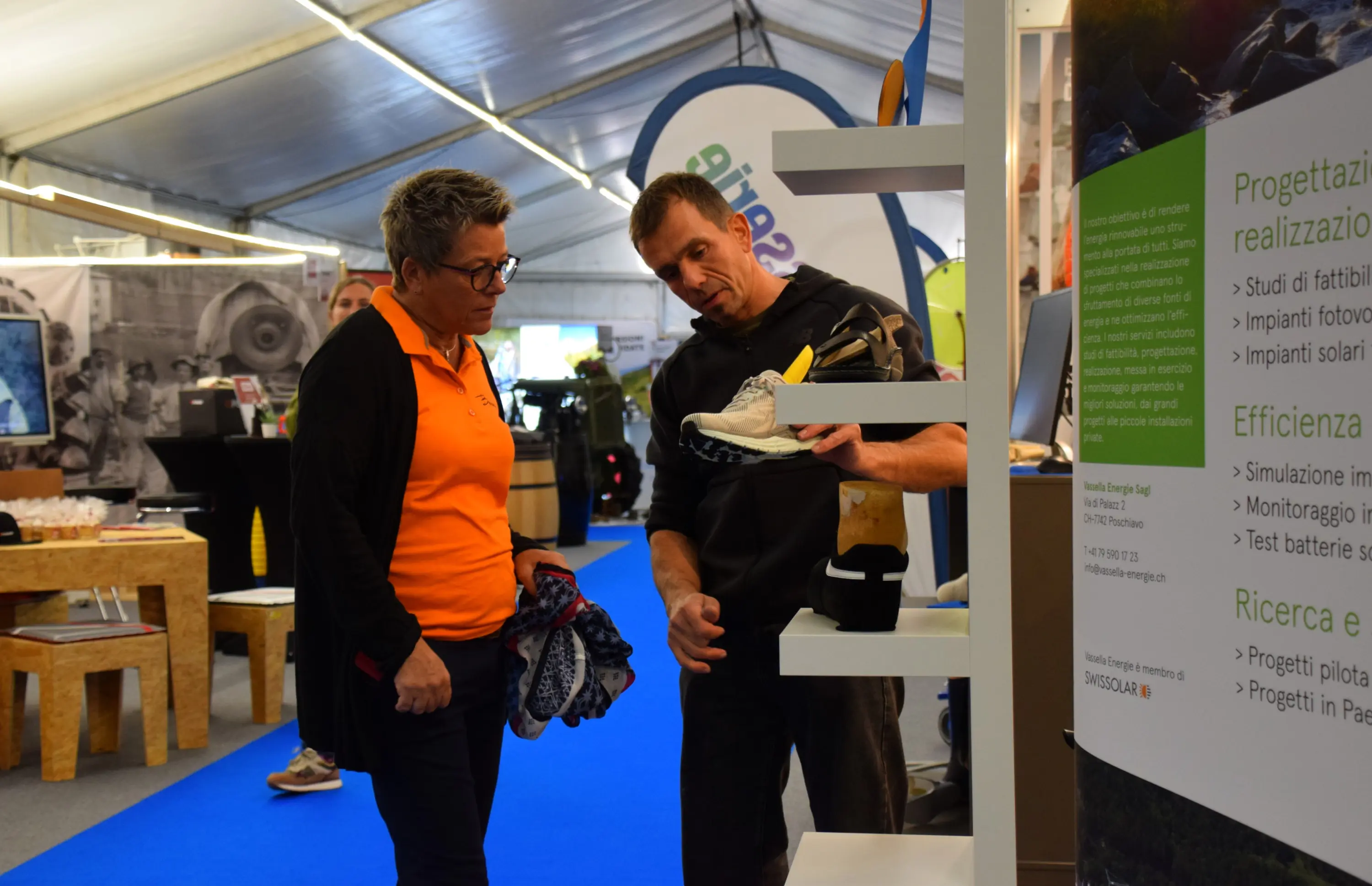 Due persone in una fiera stanno discutendo. Un uomo mostra una scarpa da trekking a una donna. Entrambi sono in un ambiente di esposizione con vari stand attorno.