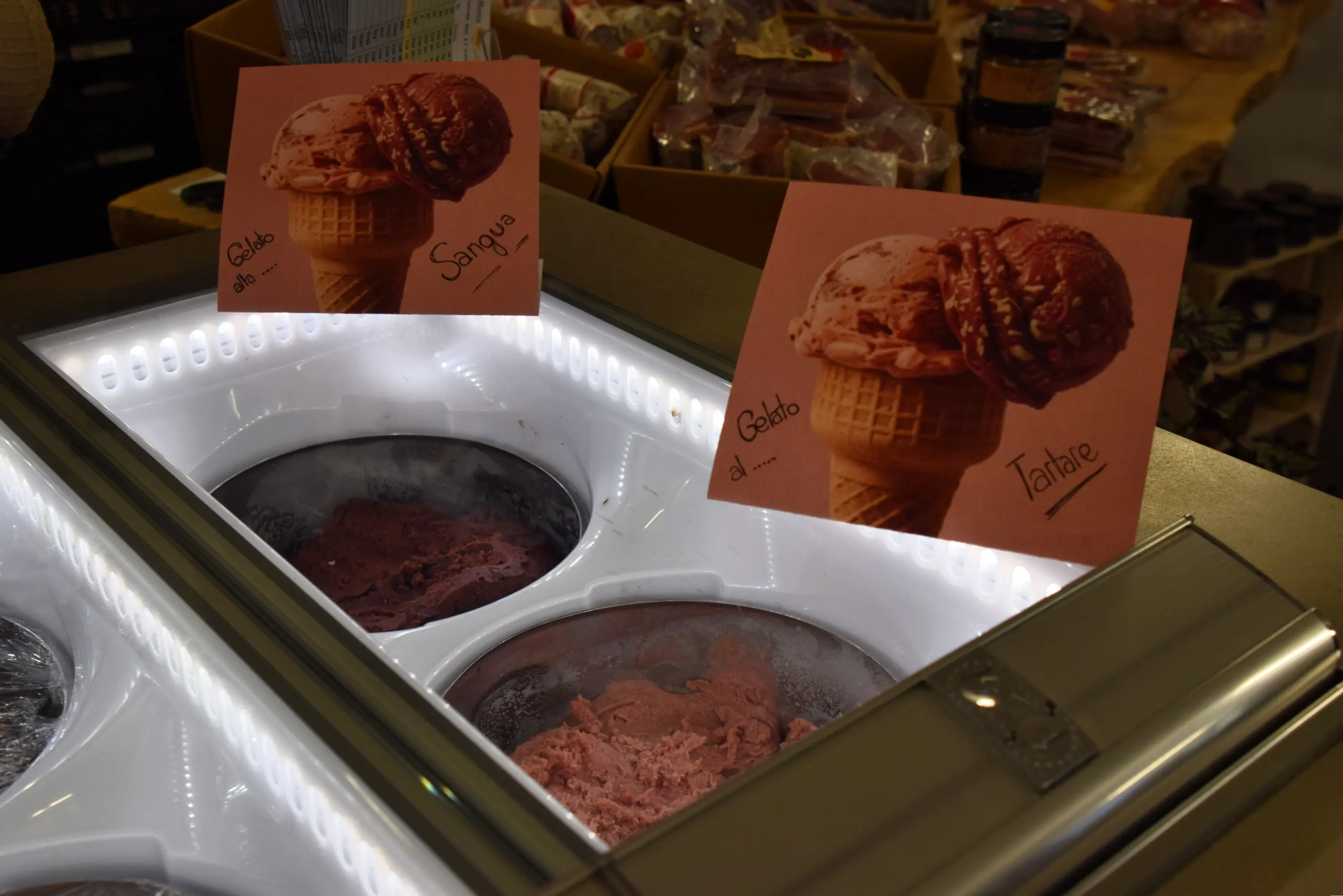 Gelato esposto in una gelateria, con cartelli che indicano due gusti: "Gelato al Sangue" e "Gelato al Tahiti". I gusti sono in contenitori di metallo.