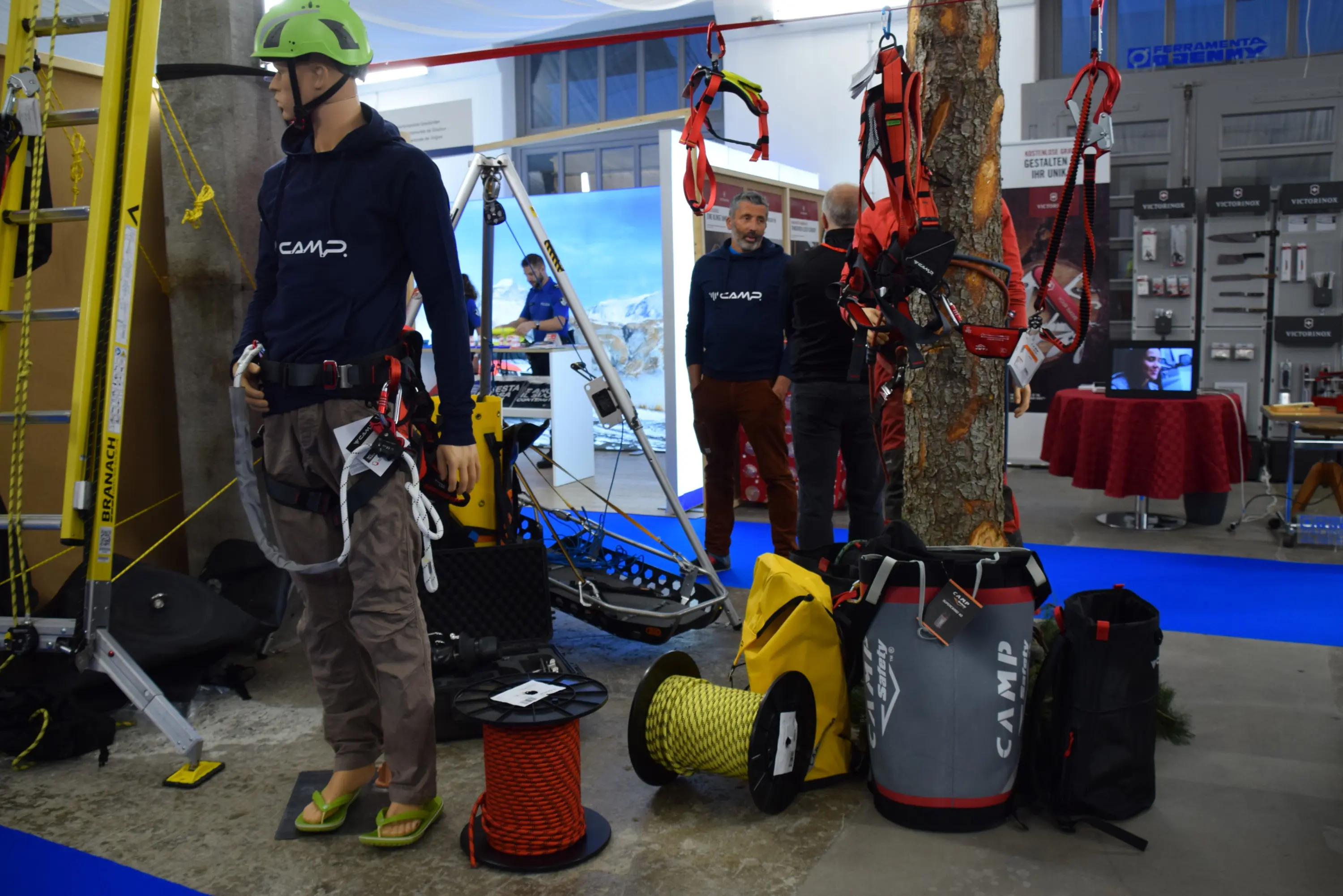 Un'esposizione con un manichino vestito in attrezzatura da arrampicata, circondato da diversi strumenti e sacchi per attrezzature. Un uomo in secondo piano osserva. Sullo sfondo, un'immagine di montagne e un'area espositiva