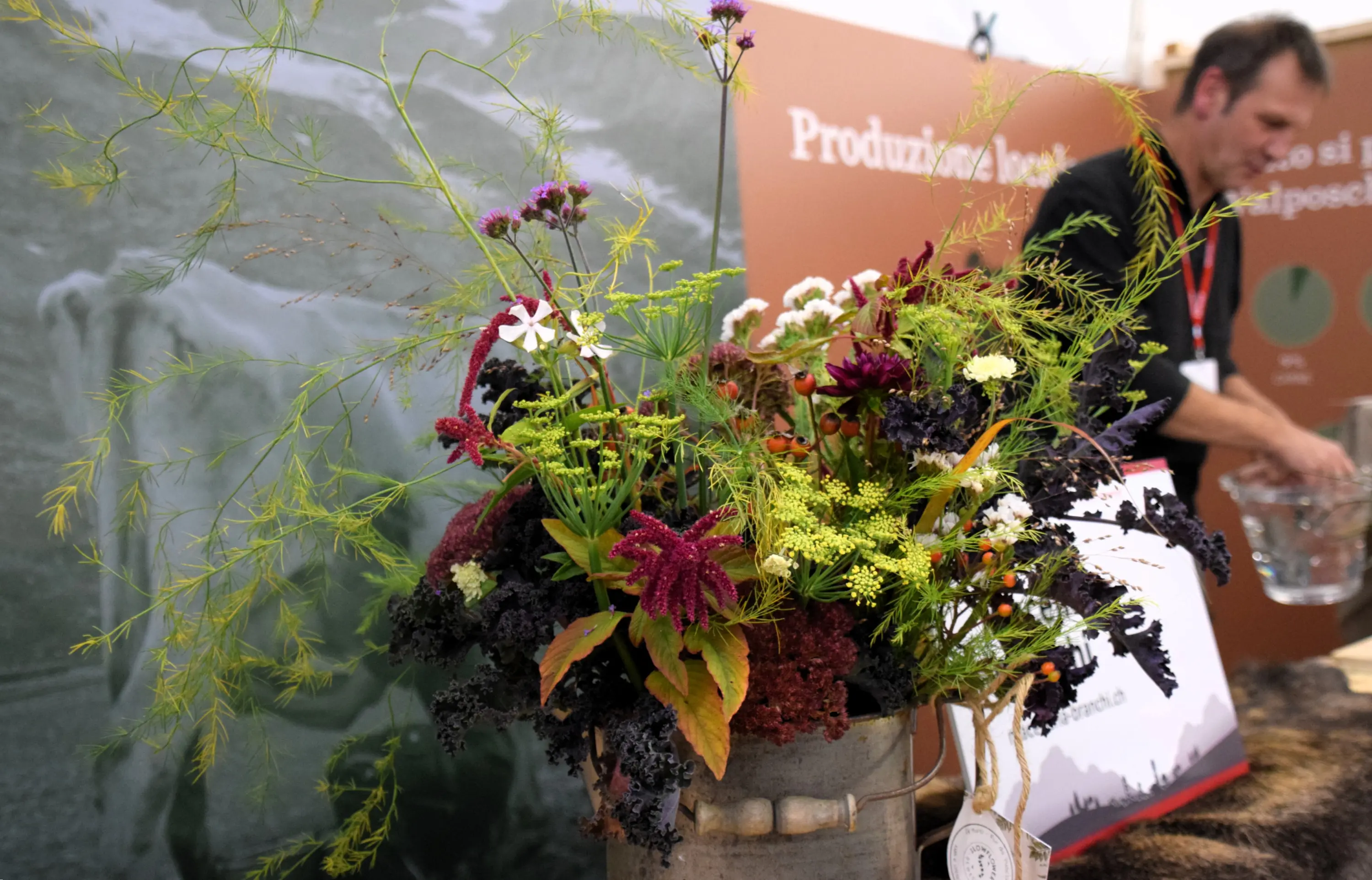 Un bouquet di fiori colorati e variopinti in un vaso, con un uomo sfocato sullo sfondo che sta preparando qualcosa. La scena è all'interno di un evento con fondali decorativi.