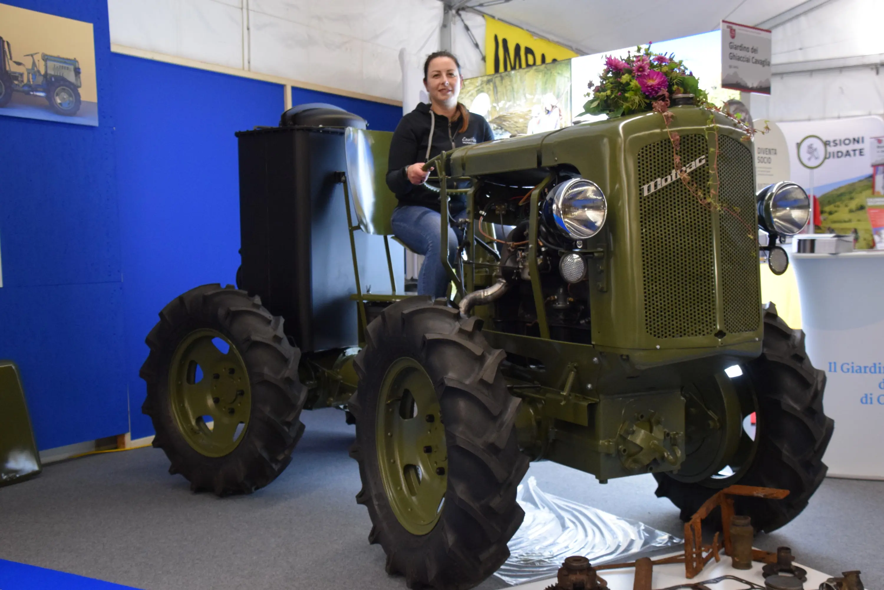 Una donna sorride mentre si trova su un trattore verde vintage, decorato con fiori, all'interno di un tendone espositivo. Sullo sfondo, si vedono pannelli informativi e altre attrezzature agricole.