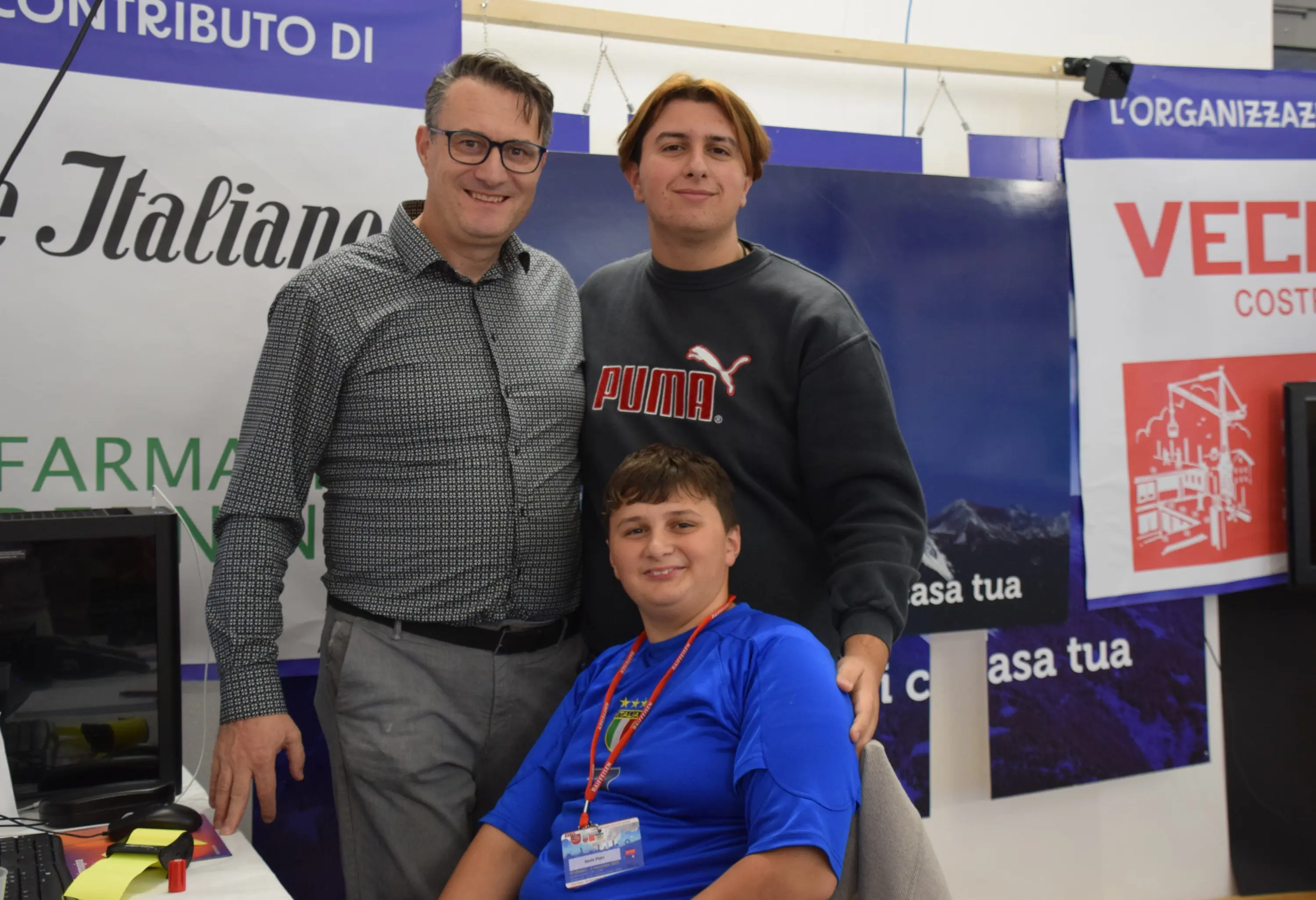 Tre persone in un ambiente di lavoro. Due uomini adulte, uno in camicia con occhiali e l'altro con una maglietta scura, sorridono accanto a un ragazzo seduto davanti a un computer. Sullo sfondo si vedono banner con scritte.