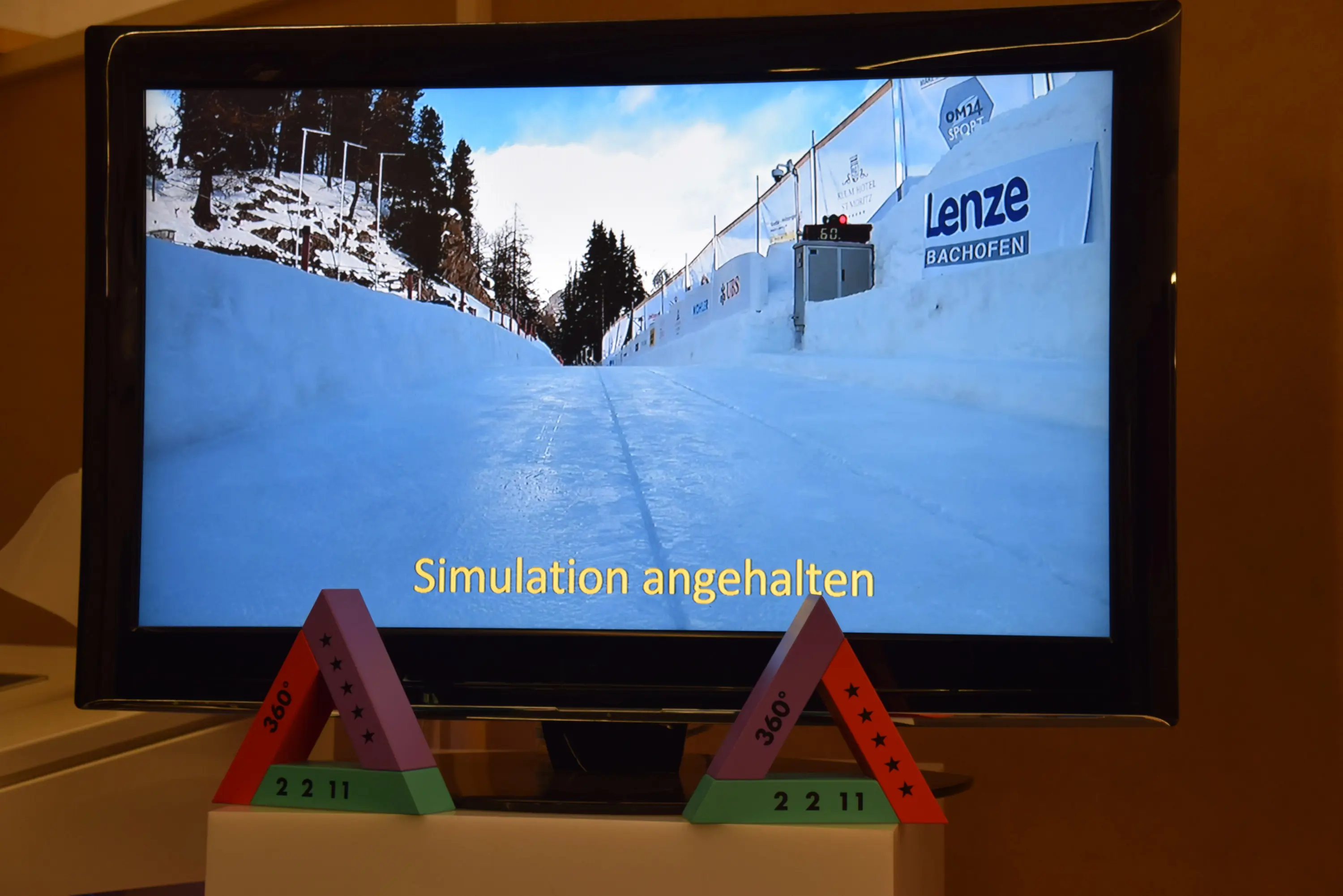 Uno schermo che mostra un percorso ghiacciato, con la scritta "Simulation angehalten" in basso. Davanti allo schermo, ci sono modelli geometrici a forma di piramide con numeri e stelle.