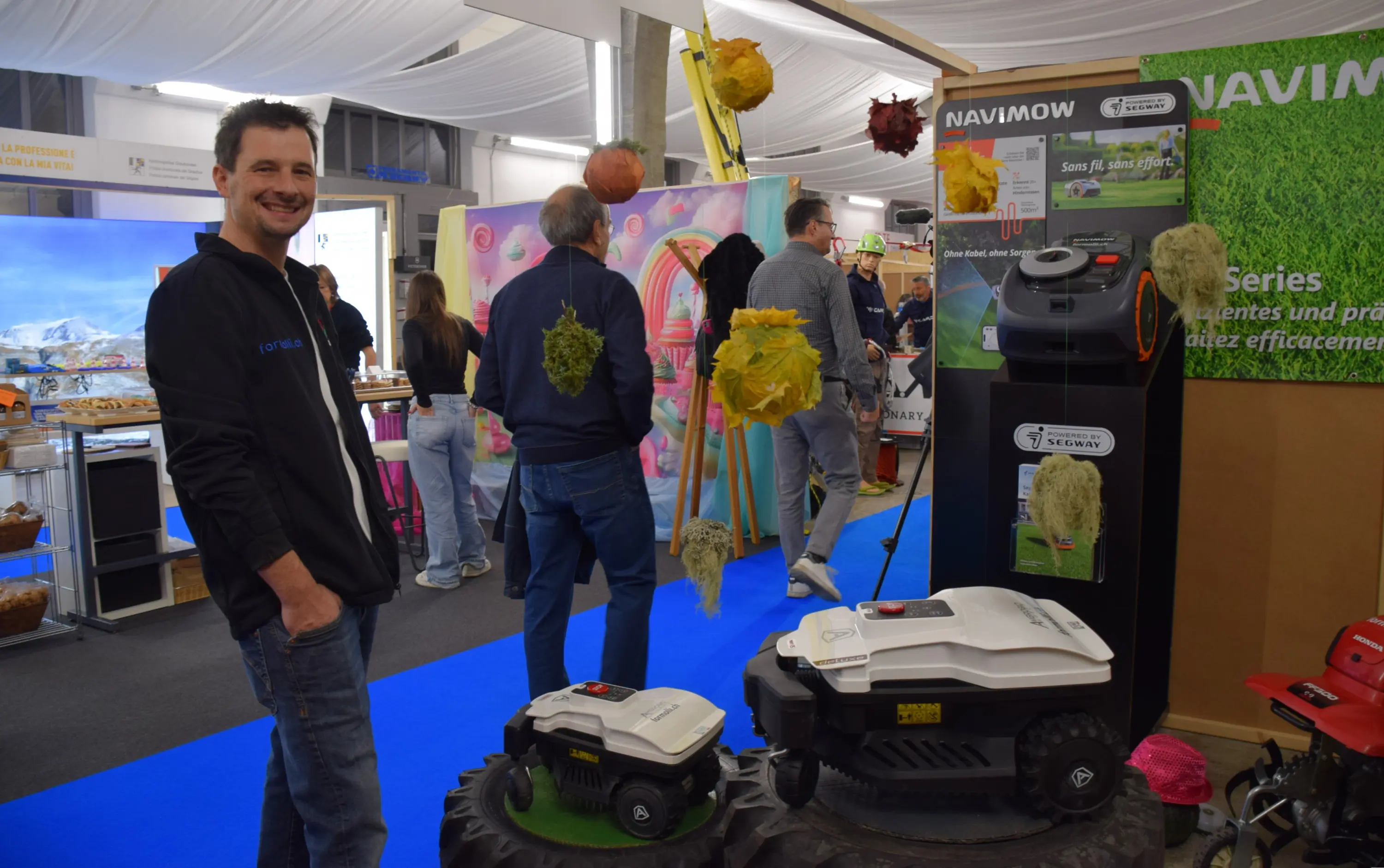 Un uomo sorridente si trova accanto a due robot tosaerba in un'esposizione, circondato da visitatori e stand vari. Sullo sfondo, alcune decorazioni colorate pendono dal soffitto.