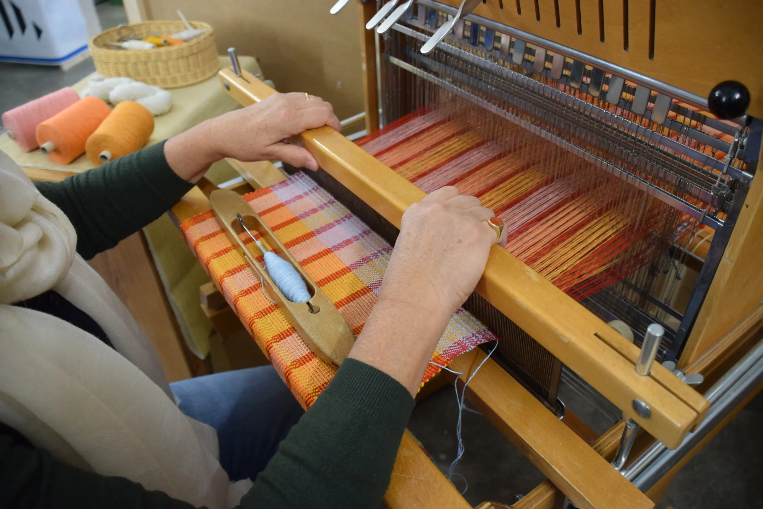 Due mani al lavoro su un telaio, mentre tessono un motivo a strisce multicolore. Lo sfondo è decorato con fili colorati di cotone e una cesta.