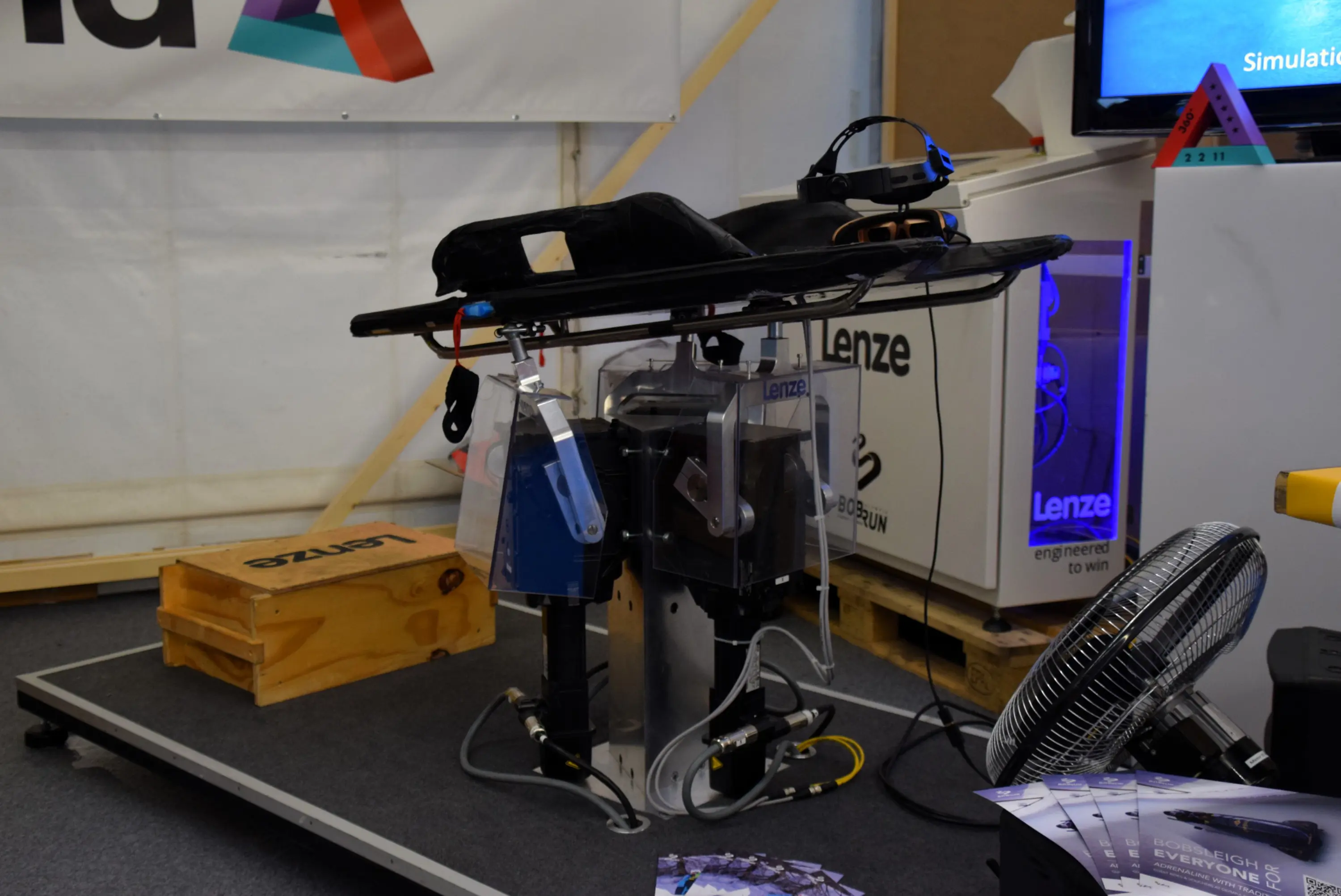 Robot industriale su una piattaforma, con componenti visibili e collegamenti a tubi. Sullo sfondo, attrezzature e materiale promozionale. La scena è allestita in un ambiente fieristico.