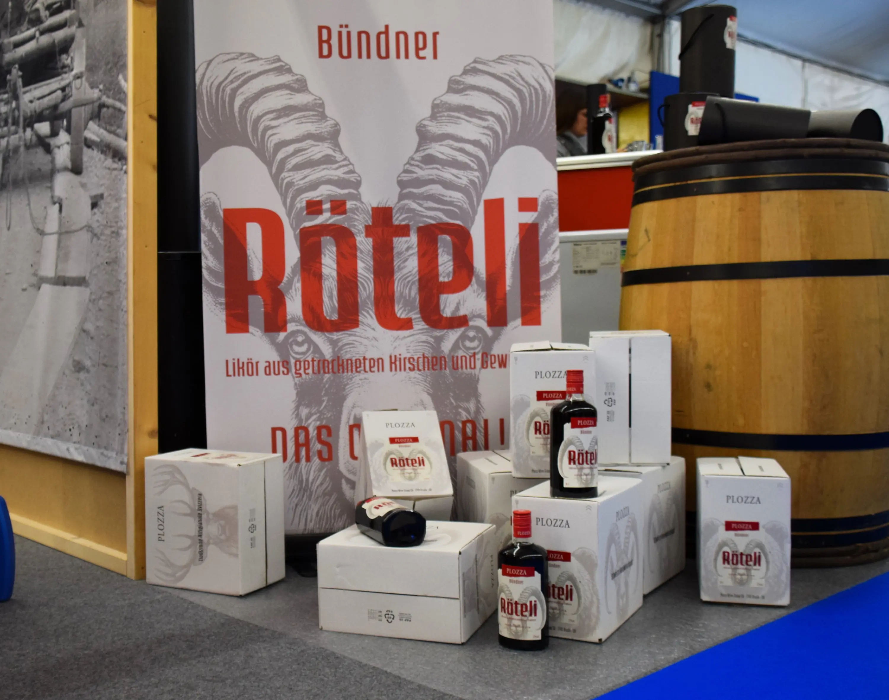 Scatole di liquore Röteli in un'area espositiva, con un grande cartellone sullo sfondo che ne promuove il marchio. Un grande barile di legno è visibile a sinistra.