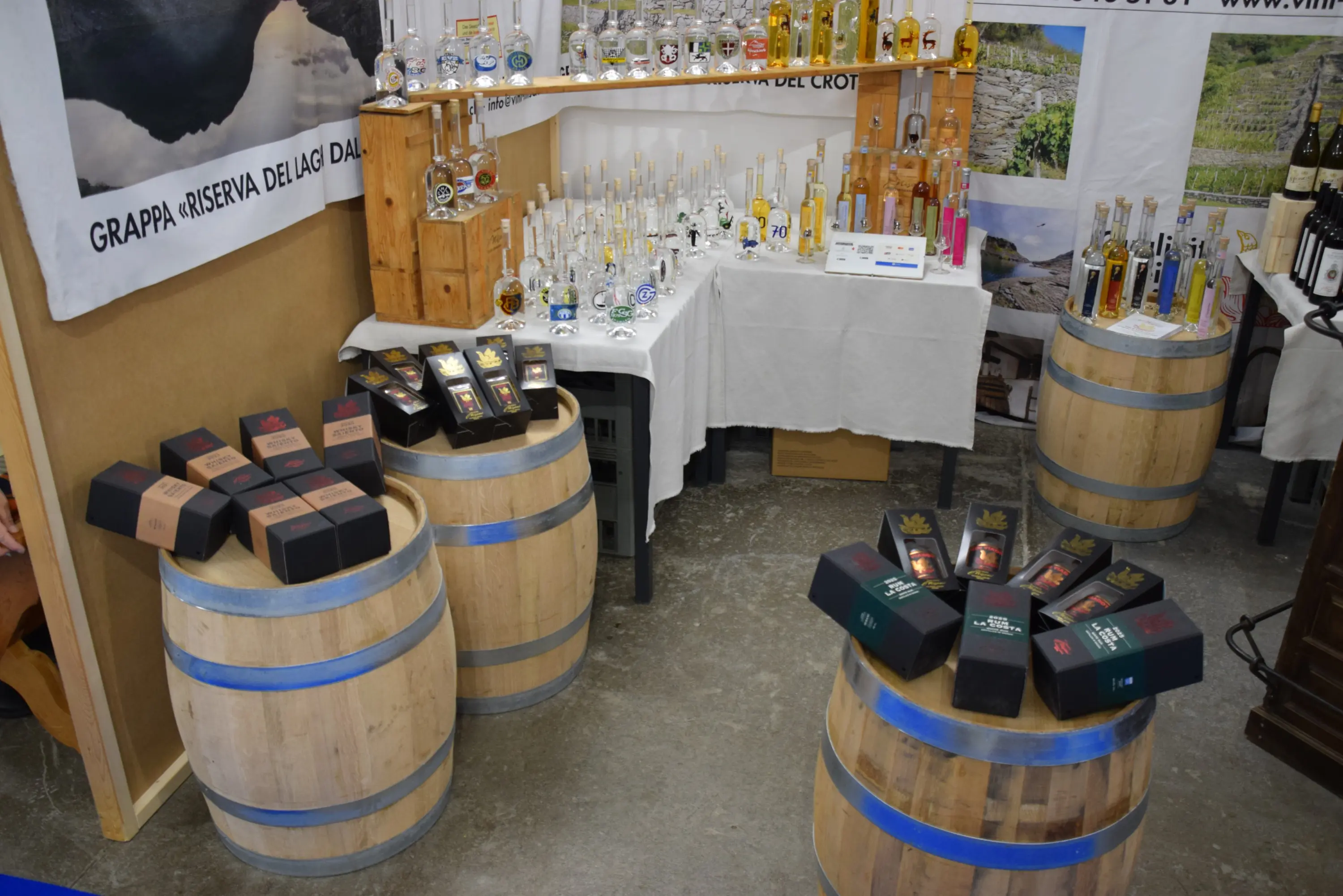 Vari tipi di grappa e liquori esposti su tavoli e barili di legno. Bottiglie di diverse forme e colori sono disposte in modo ordinato. Alcuni pacchetti regalo sono in mostra in primo piano.