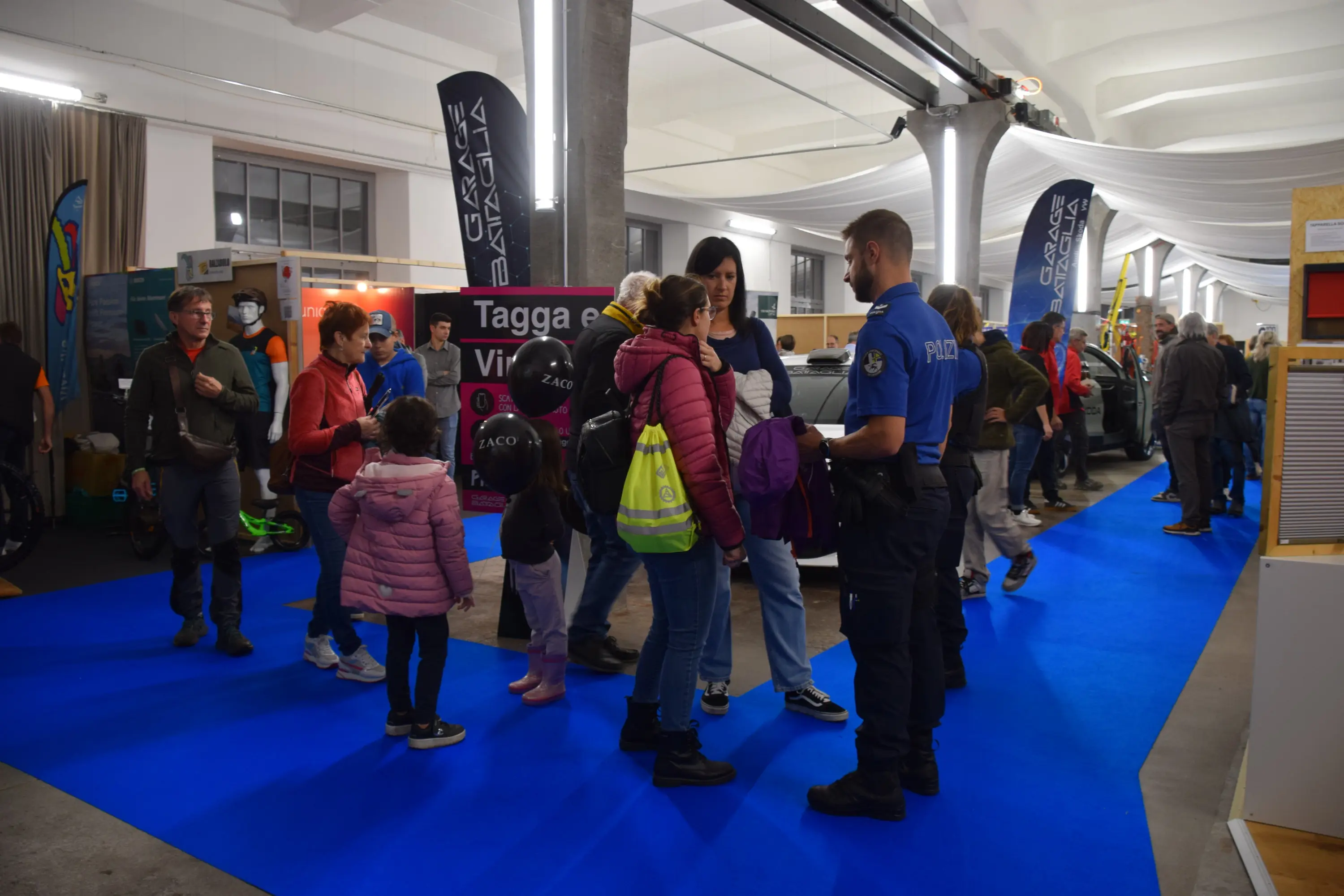 Una fiera con visitatori che si trovano tra stand. Alcuni poliziotti sono in conversazione con una famiglia. Il pavimento è coperto da un tappeto blu. Diverse bandiere e cartelli decorano l'area.