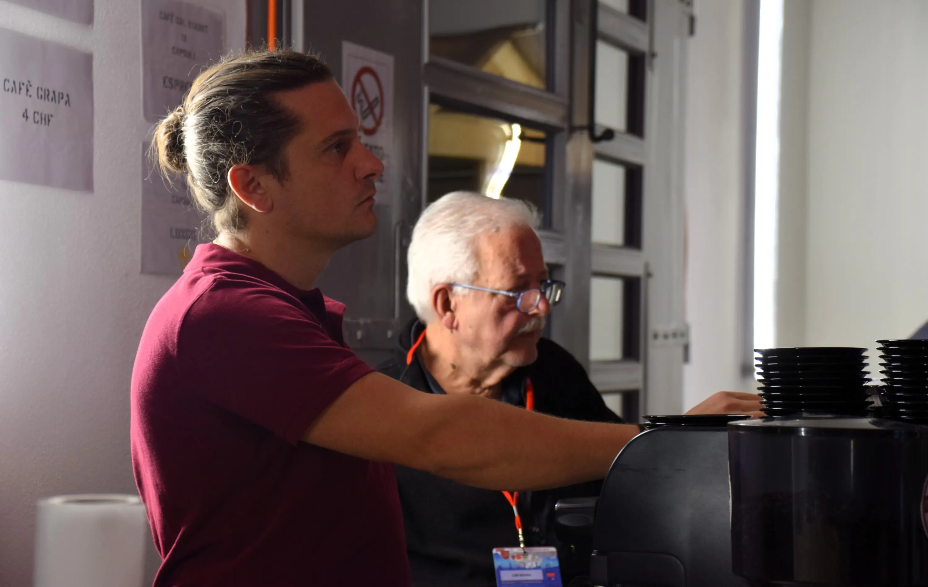 Un giovane barista con capelli raccolti prepara caffè, mentre un uomo anziano con occhiali osserva attentamente. Sullo sfondo si vedono poster e tazze.