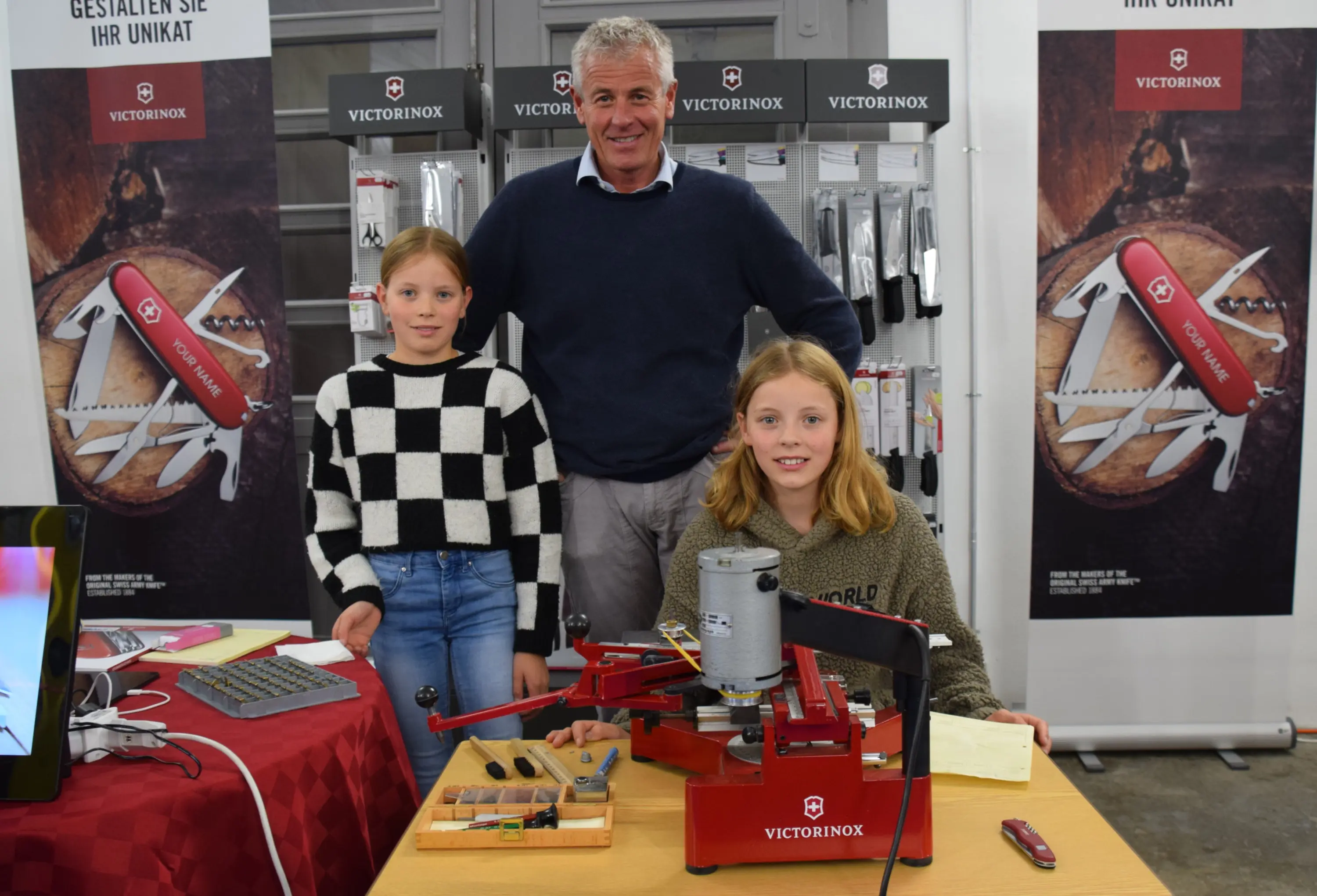 Due ragazze sorridenti si trovano davanti a un tavolo con attrezzature di personalizzazione. Un uomo adulto sta dietro di loro, tutti e tre posano per la foto. Sullo sfondo si intravedono i banner del marchio Victorinox.