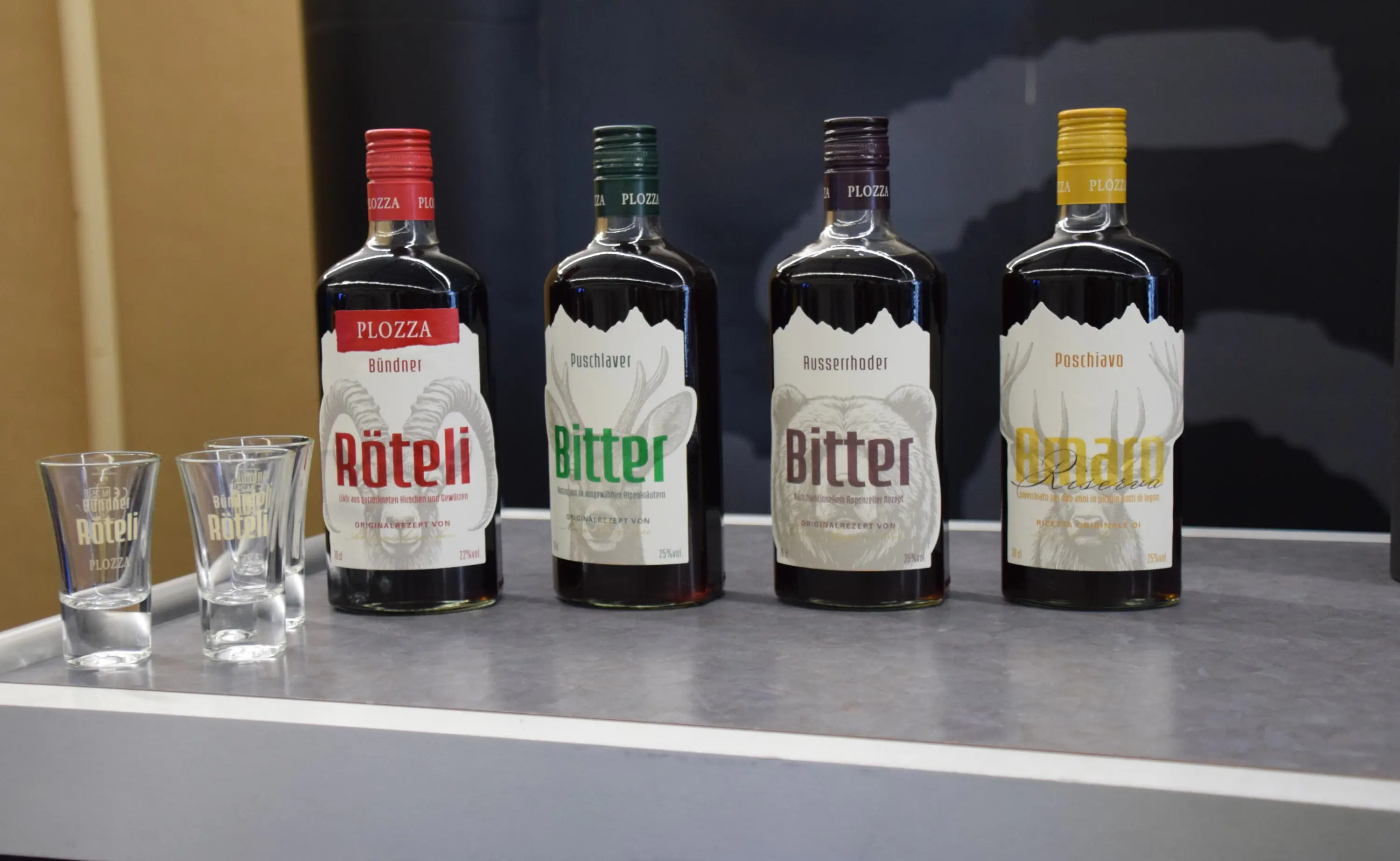 Quattro bottiglie di liquore su un tavolo, etichettate Röteli, Bitter e Oro. Due bicchieri trasparenti accanto alle bottiglie. Sfondo neutro.