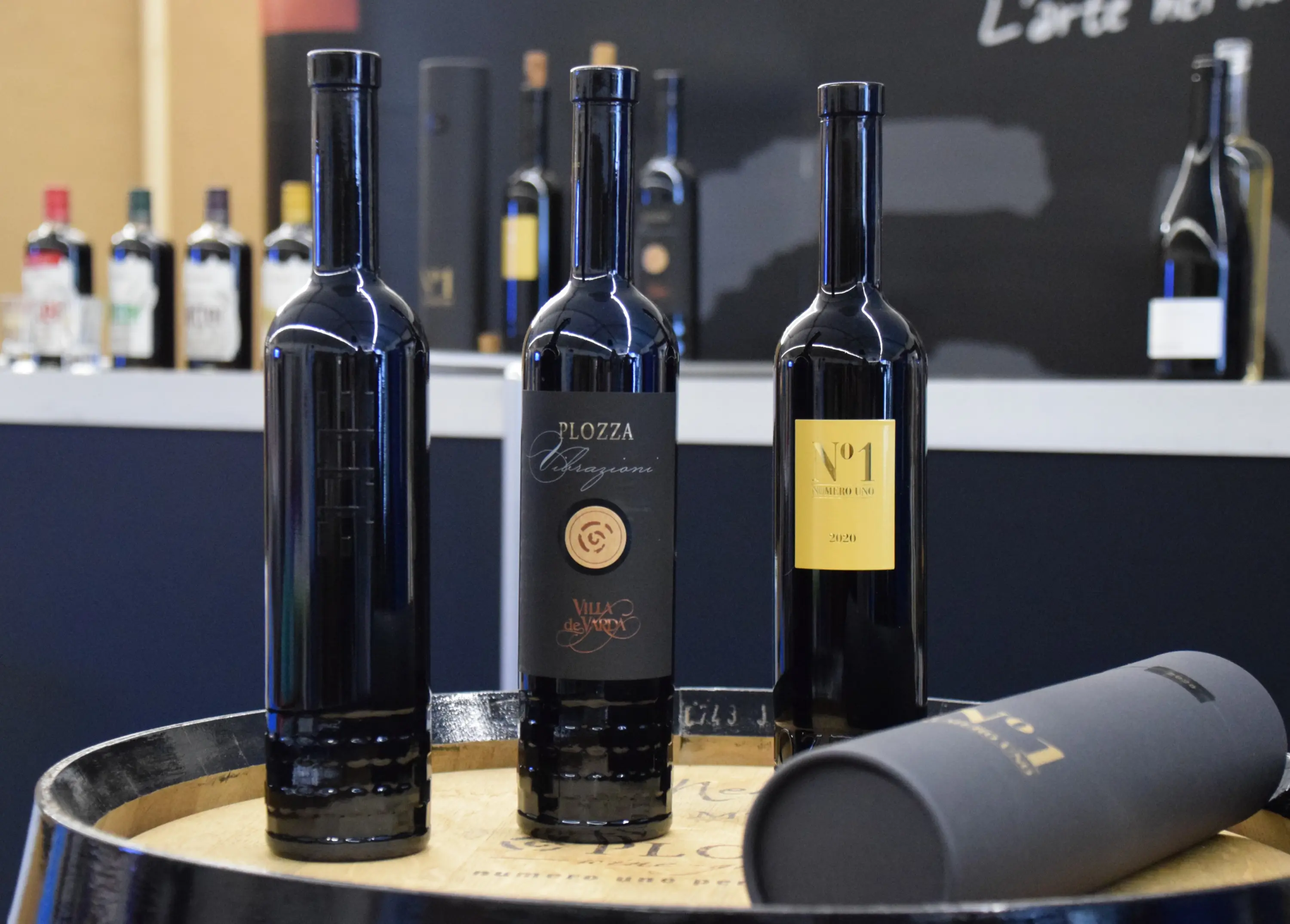 Tre bottiglie di vino su un barile di legno. Le etichette mostrano diversi design: una bottiglia ha un'etichetta dorata con il numero 1, un'altra è nera con un logo centrale e l'ultima è più semplice con il nome "Plo