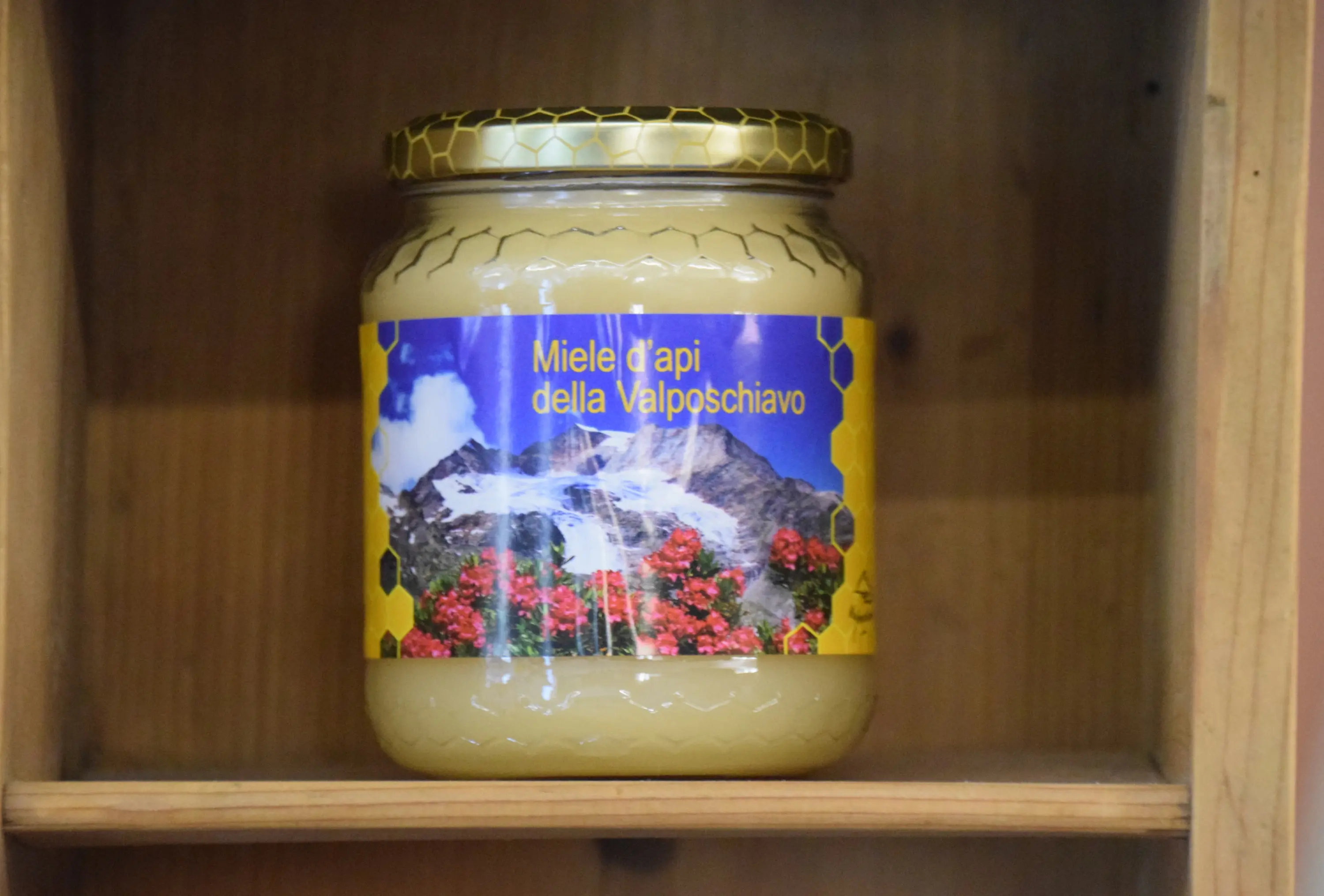 Barattolo di miele con etichetta "Mielle d'api della Valposchiavo", decorato da immagini di fiori e montagne. È collocato su uno scaffale di legno.