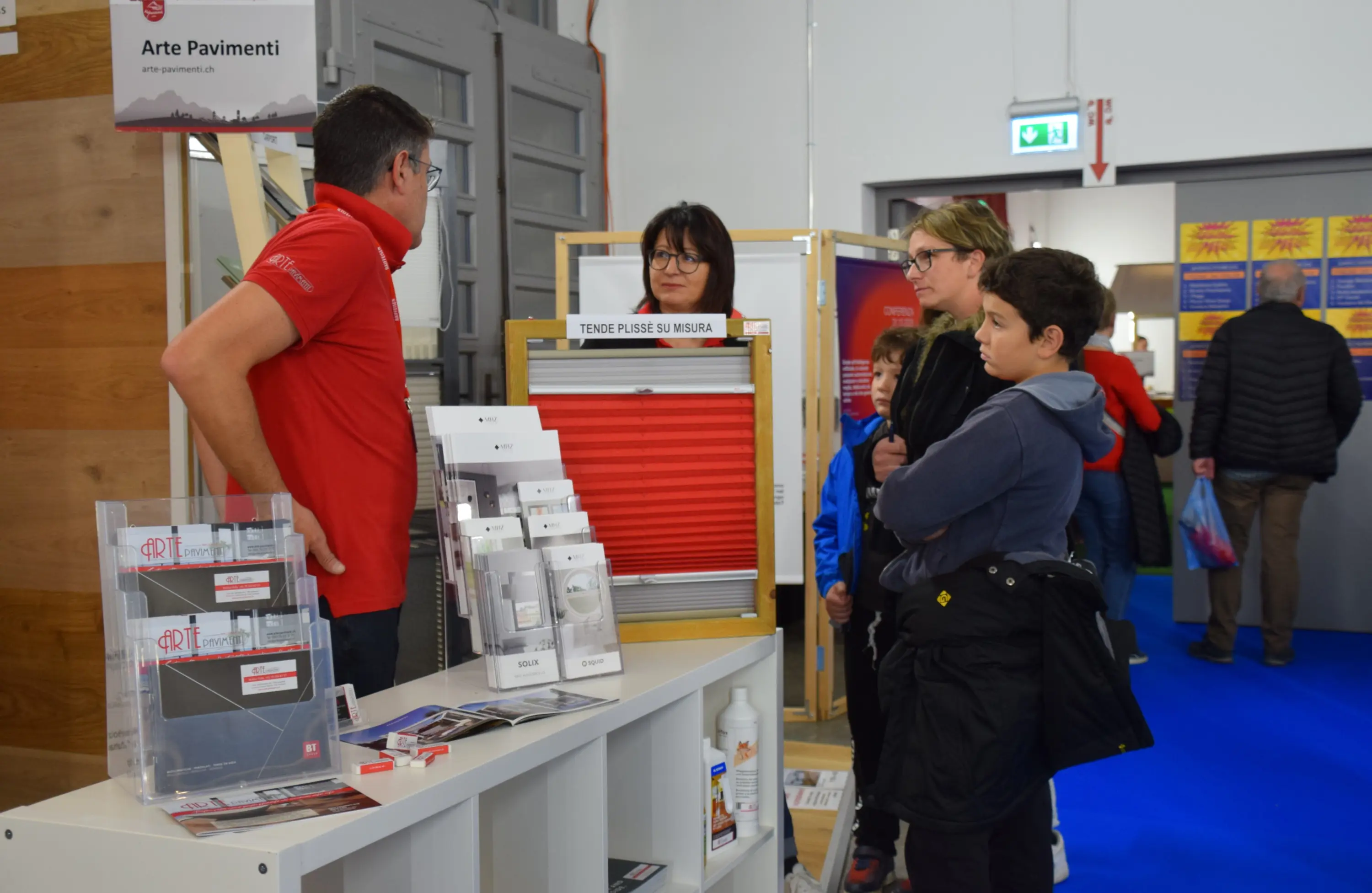 Un uomo in maglietta rossa spiega un prodotto a una donna e un ragazzo in un evento fieristico. Al tavolo sono presenti materiali informativi e campioni. Sullo sfondo, altre persone sono visibili in un ambiente espositivo.