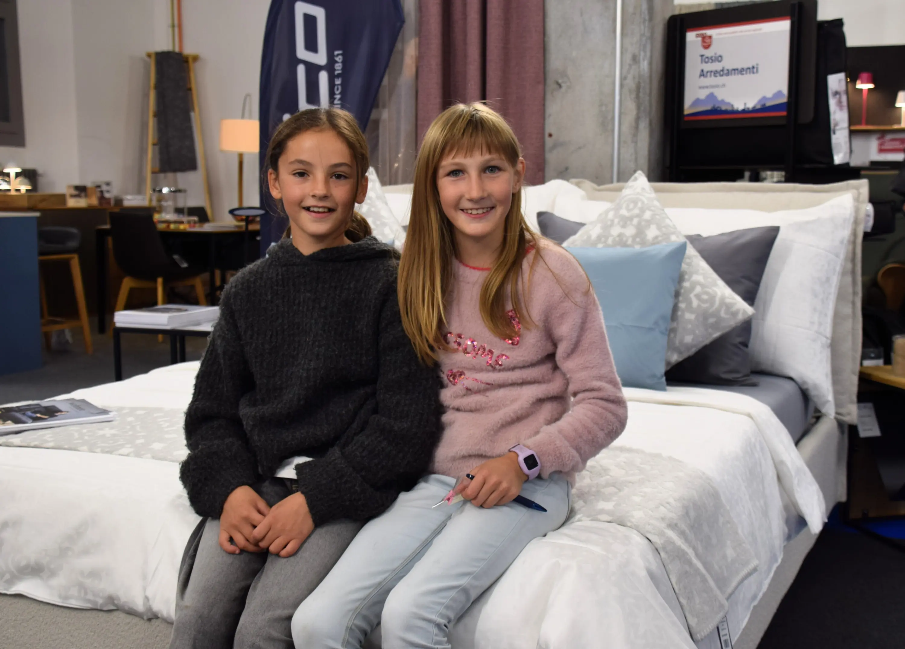 Due ragazze sorridenti siedono su un letto in un negozio di arredo. Una indossa un maglione nero, mentre l'altra ha un maglione rosa. Sullo sfondo si intravedono diversi mobili e decorazioni.