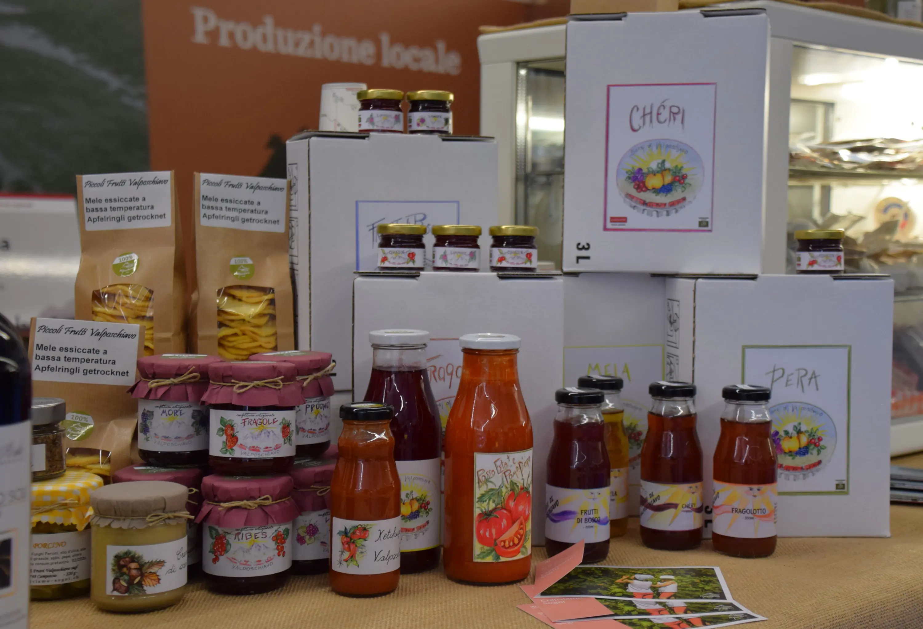 Vari prodotti alimentari locali in esposizione, tra cui conserve, succhi e marmellate, tutti etichettati con nomi come "Ciliegia", "Mela" e "Pera". I contenitori sono disposti su un tavolo con una tovaglia beige.