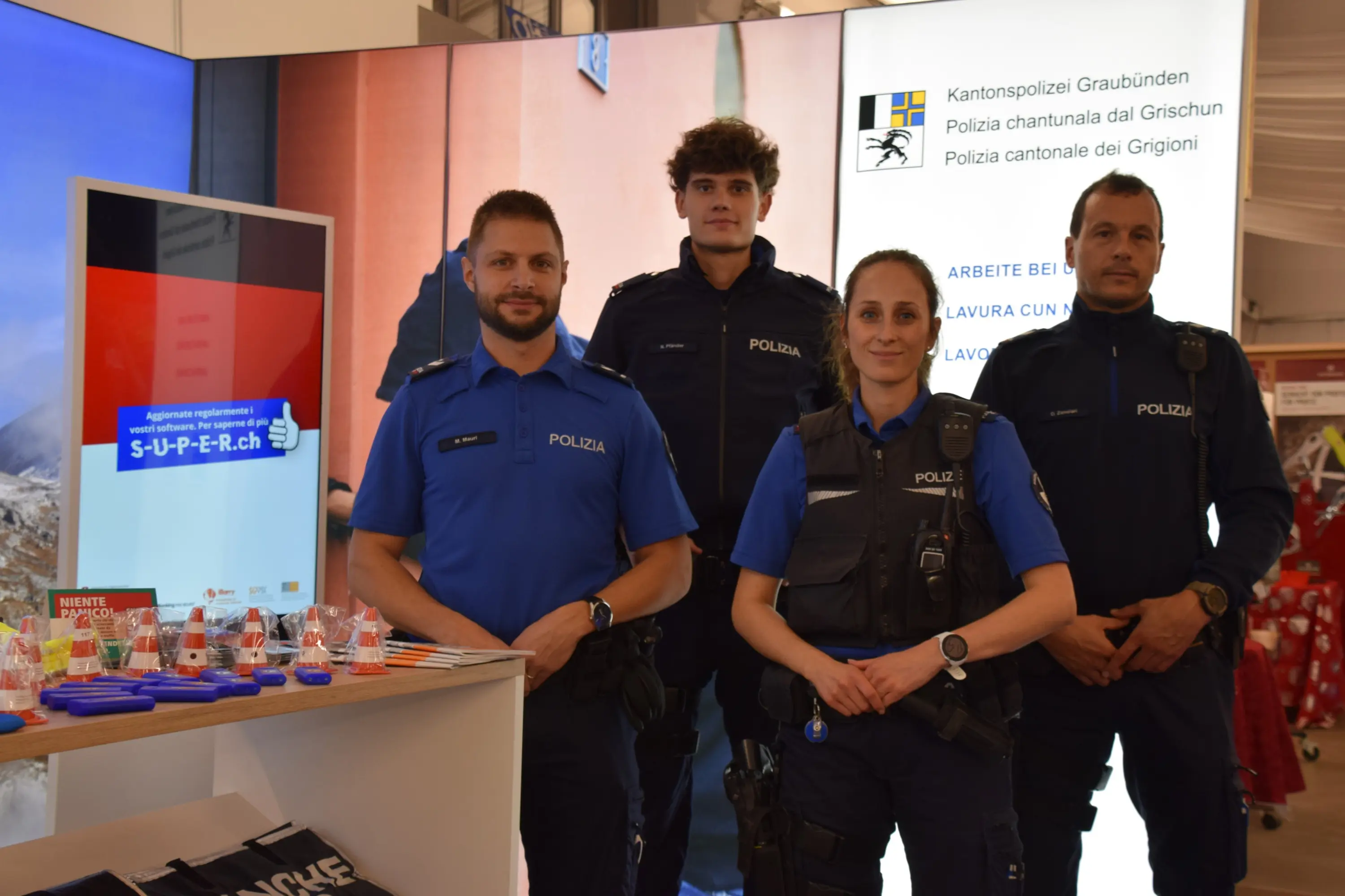 Quattro membri della polizia cantonale dei Grigioni, in uniforme, sorridono davanti a uno stand promozionale. Sullo sfondo, un display evidenzia informazioni sulla loro attività.