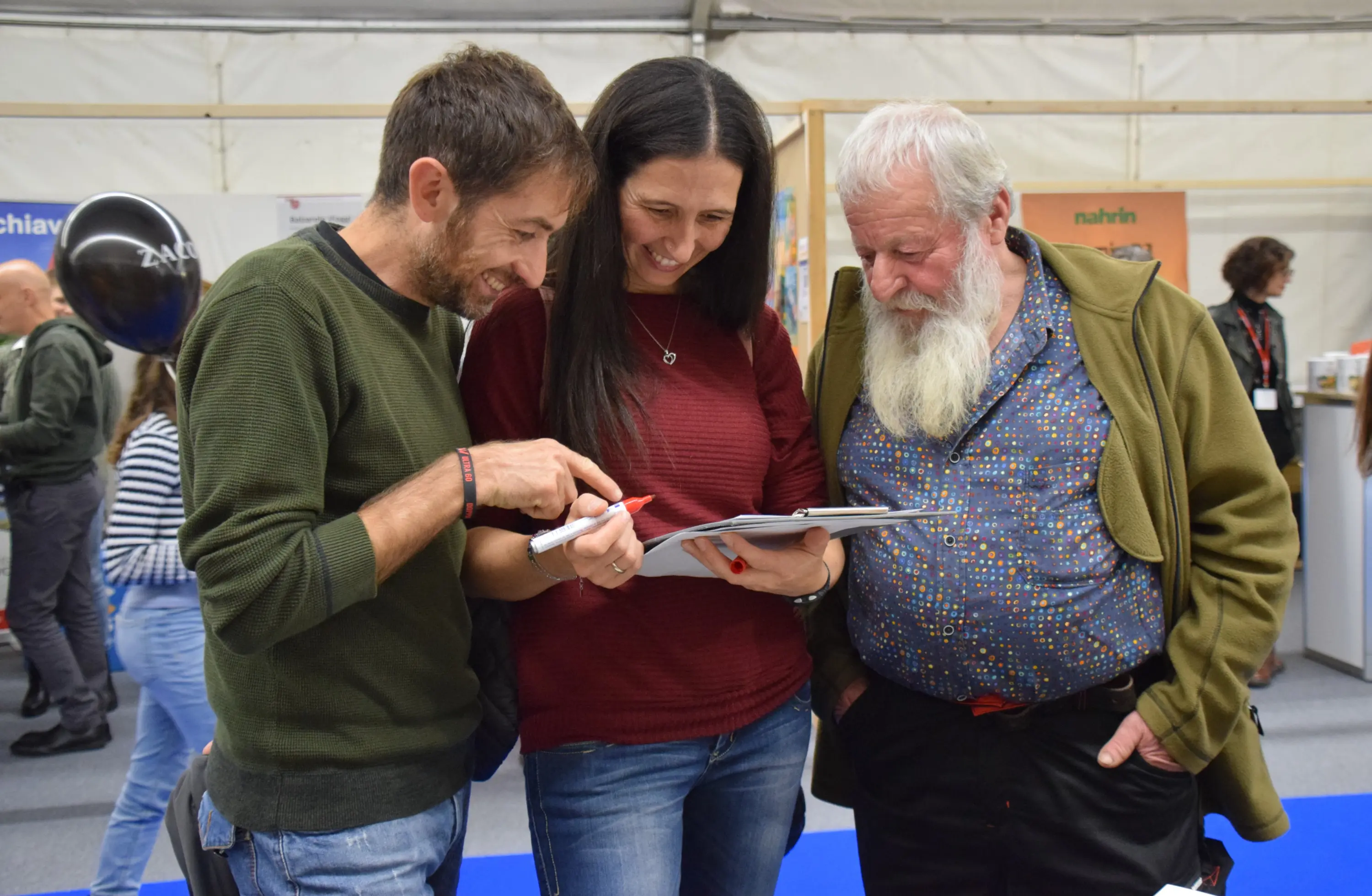 Tre persone stanno esaminando un documento insieme, sorridendo e conversando. Un uomo a sinistra ha i capelli corti e brizzolati, una donna al centro indossa un maglione rosso, mentre un uomo a destra ha una lunga barba bianca e un